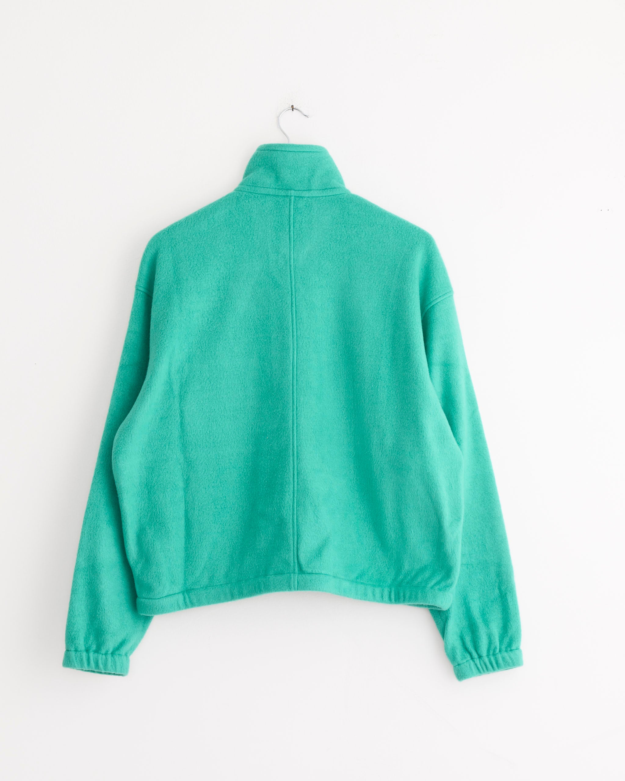 The Auralee Pure Silk Half Zip Pullover in Jade Green, featuring long sleeves and a stand-up collar, is displayed from the back on a simple hook against a plain white wall.
