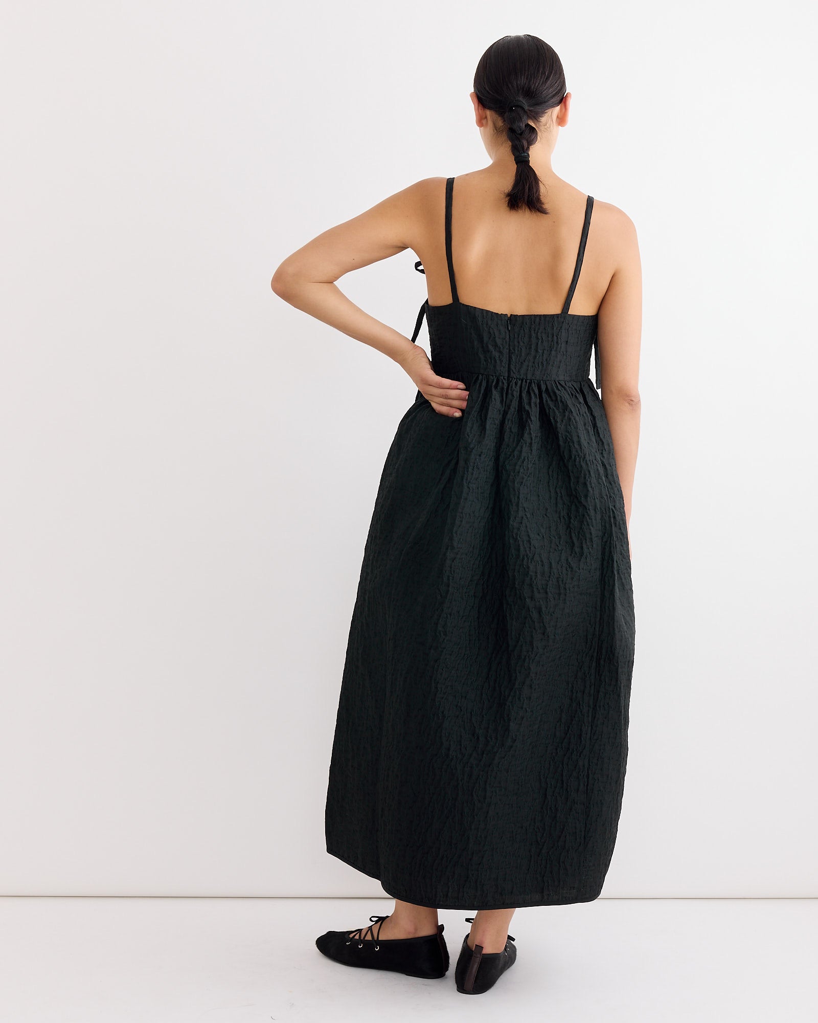 A person with dark hair in a low bun models the Cecilie Bahnsen Ambra Dress in Black, a long textured dress with thin straps, paired with black flats, standing with their back to the camera against a white background.