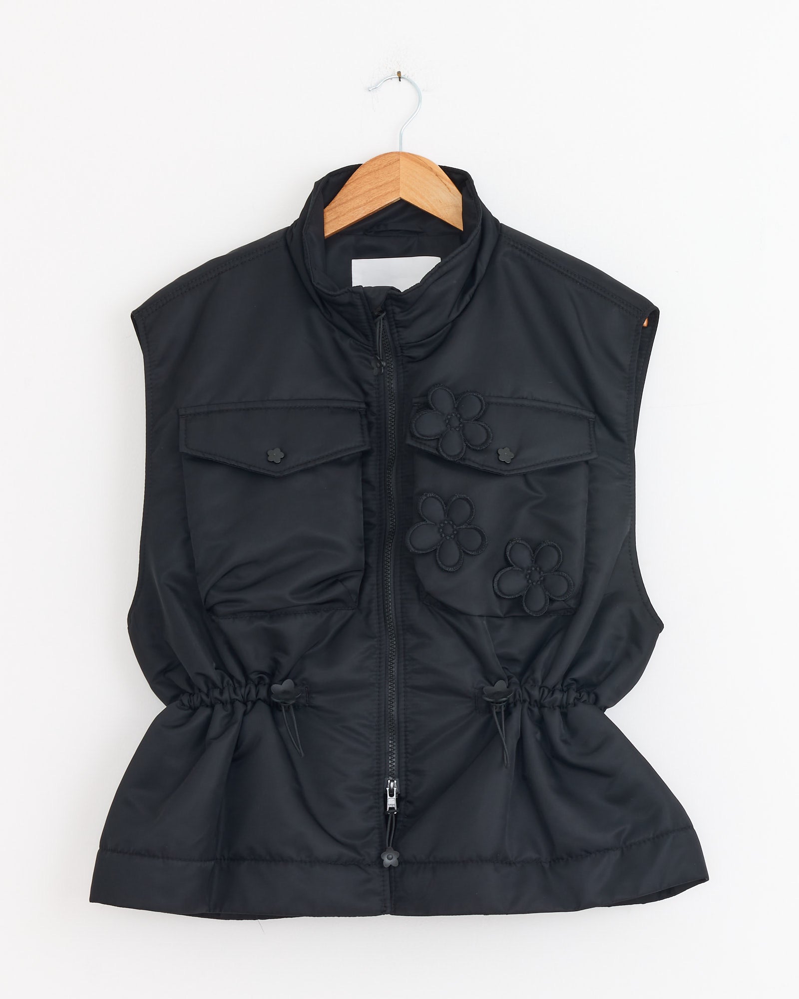 The Cecilie Bahnsen Bree Vest in Black is a technical nylon sleeveless vest with a front zipper, chest flap pockets, floral appliqués, and drawstring waist, shown on a wooden hanger against a white background.