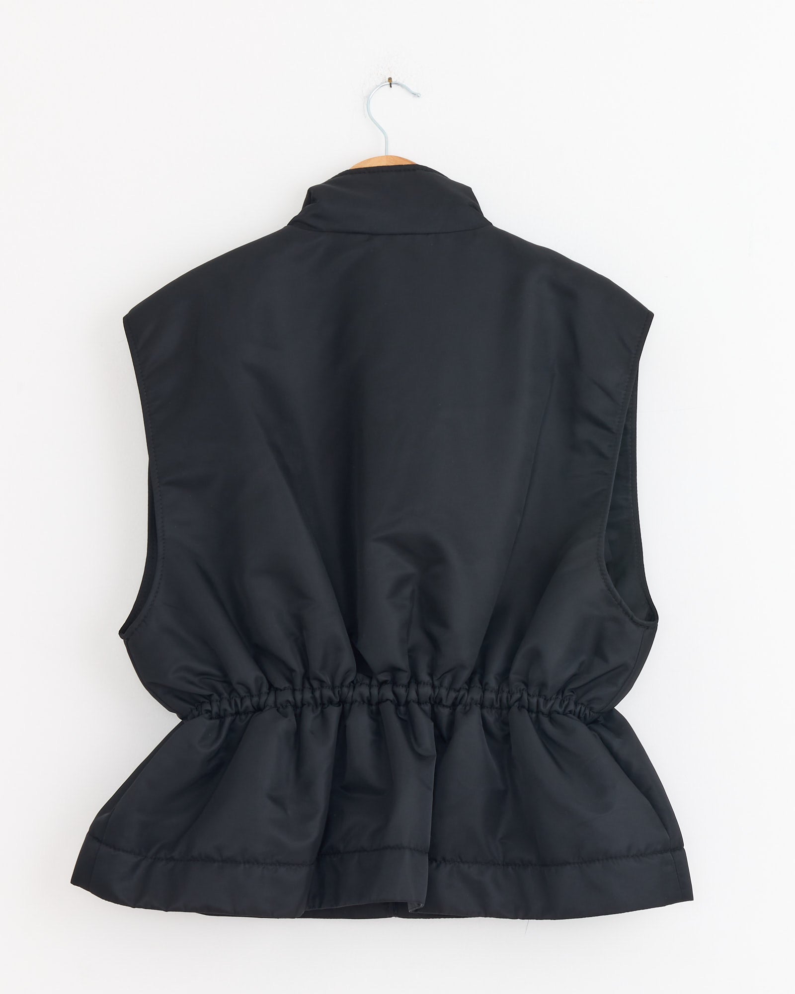 The Cecilie Bahnsen Bree Vest in Black is shown from the back, featuring technical nylon with a high collar and gathered elastic waist, displayed on a wooden hanger against a plain white background.