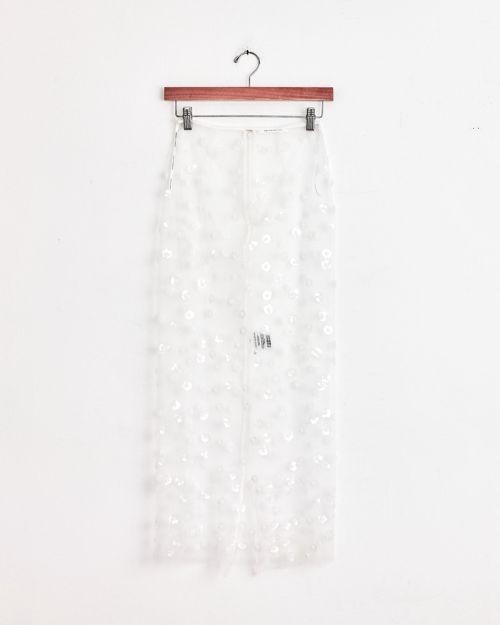 The Cecilie Bahnsen Gus Skirt in White Sequin hangs on a wooden hanger against a plain white wall.