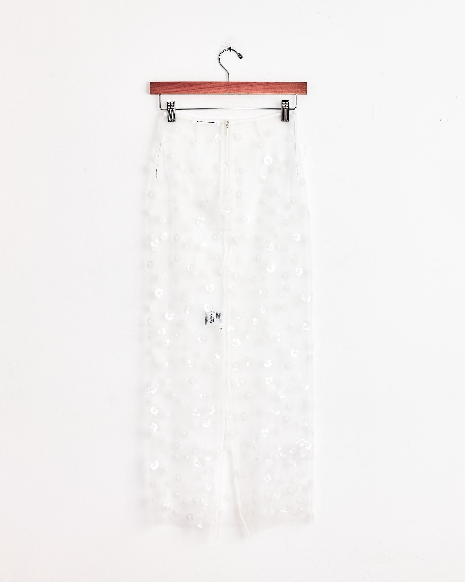 The Gus Skirt in White Sequin by Cecilie Bahnsen hangs on a wooden hanger against a plain white wall, showcasing its sheer fabric and delicate sequin embellishments for an ethereal, decorative effect.