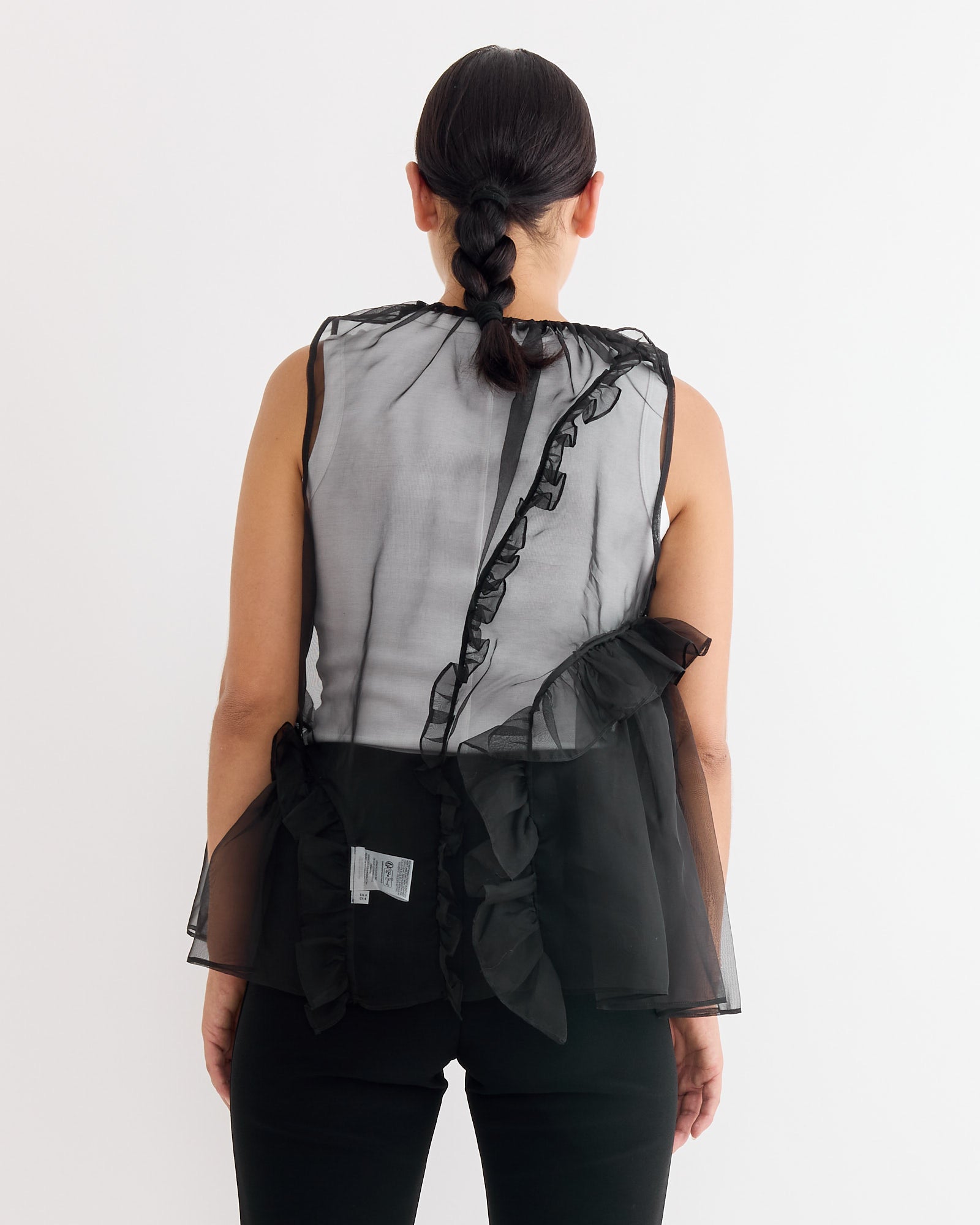 A person with dark braided hair wears the Cecilie Bahnsen Orsalina Top in Black—a sleeveless, sheer silk organza top with ruffle details—paired with black pants, standing against a plain white background.