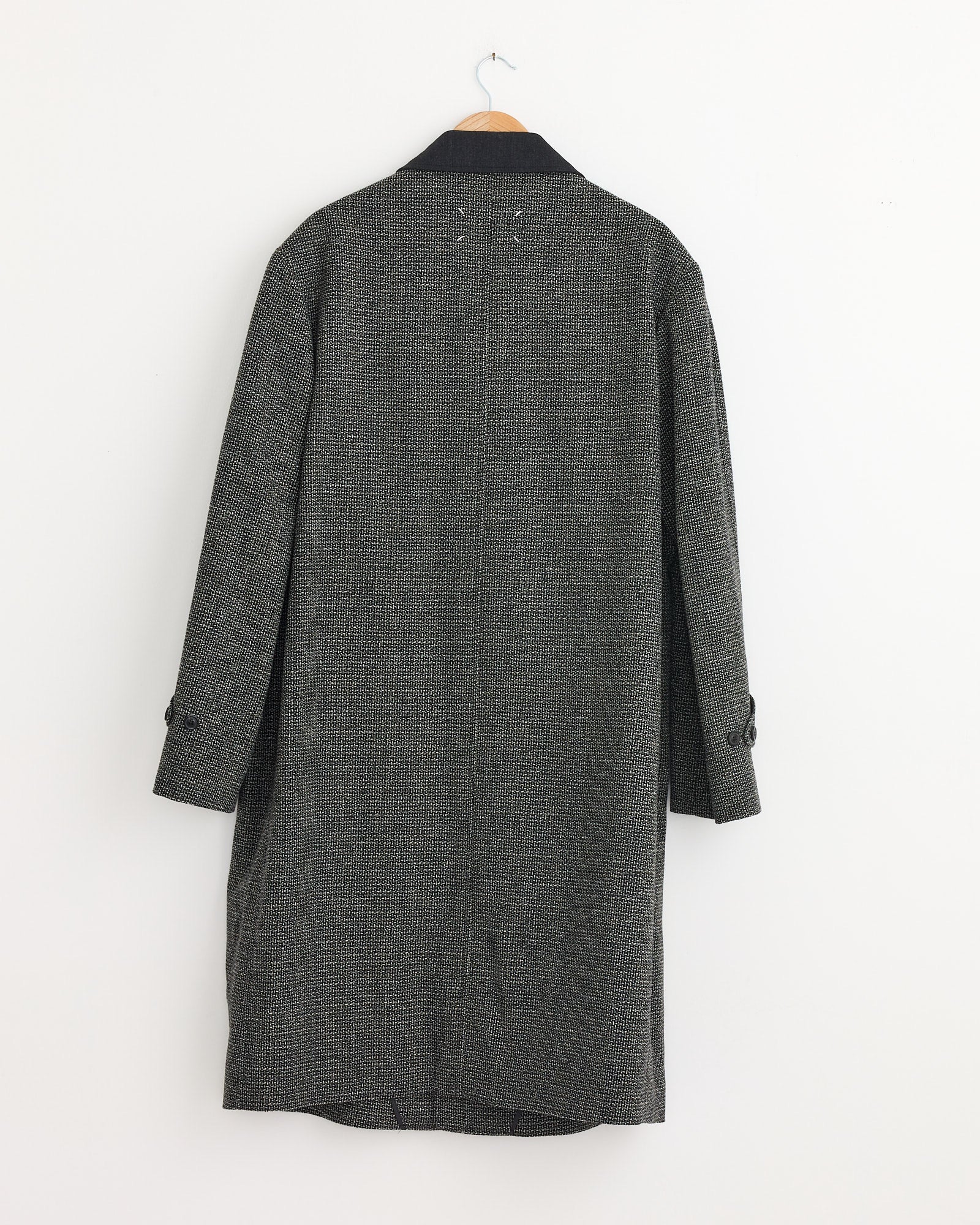 A Maison Margiela Wool Coat in Grey with black sleeve buttons hangs on a wooden hanger against a plain white wall, shown from the back.
