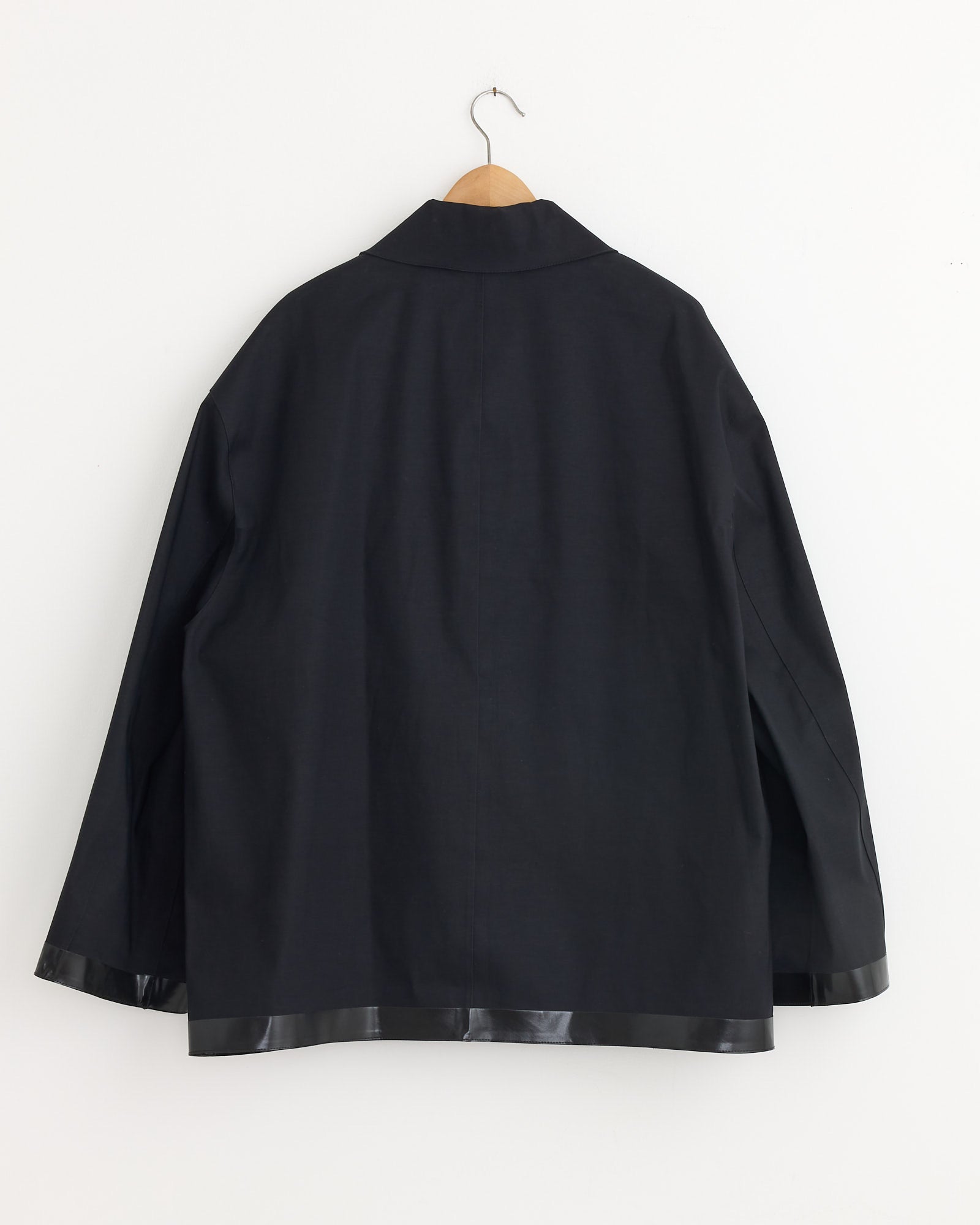 The Studio Nicholson x Mackintosh Teviot Bonded Short Coat in Black hangs on a wooden hanger against a white wall, shown from the back. It features a collar and shiny black trim along the hem and cuffs.