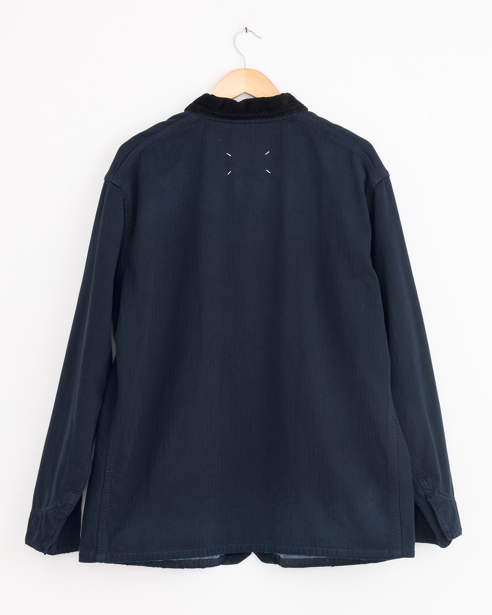 The Maison Margiela Sports Jacket in Dark Blue, featuring a black corduroy collar and two white stitches below the collar at the back, hangs on a wooden hanger against a white wall.