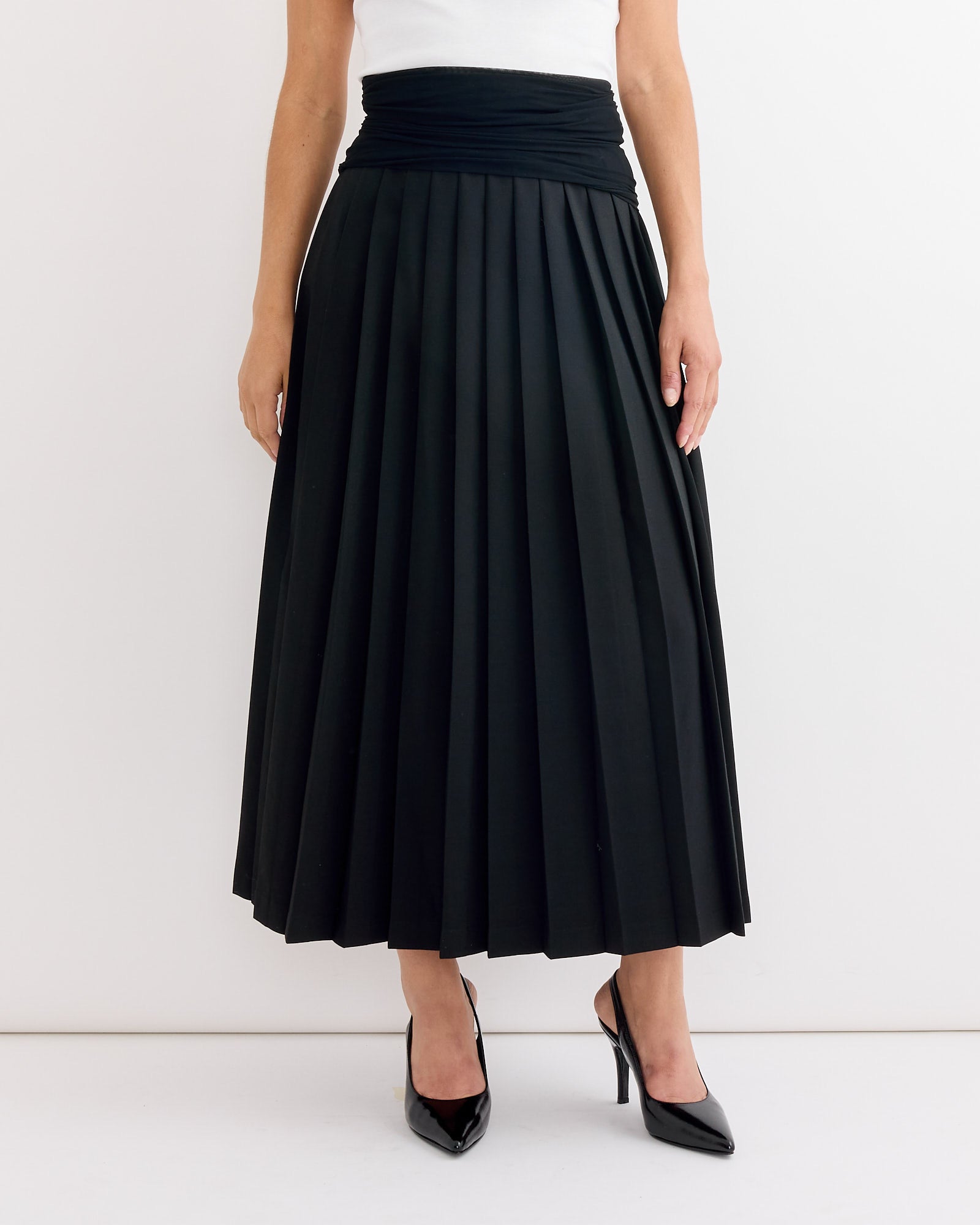A woman stands against a white background wearing a white sleeveless top, black pointed high heels, and the MM6 Maison Margiela Long Skirt in Black with structured pleats.