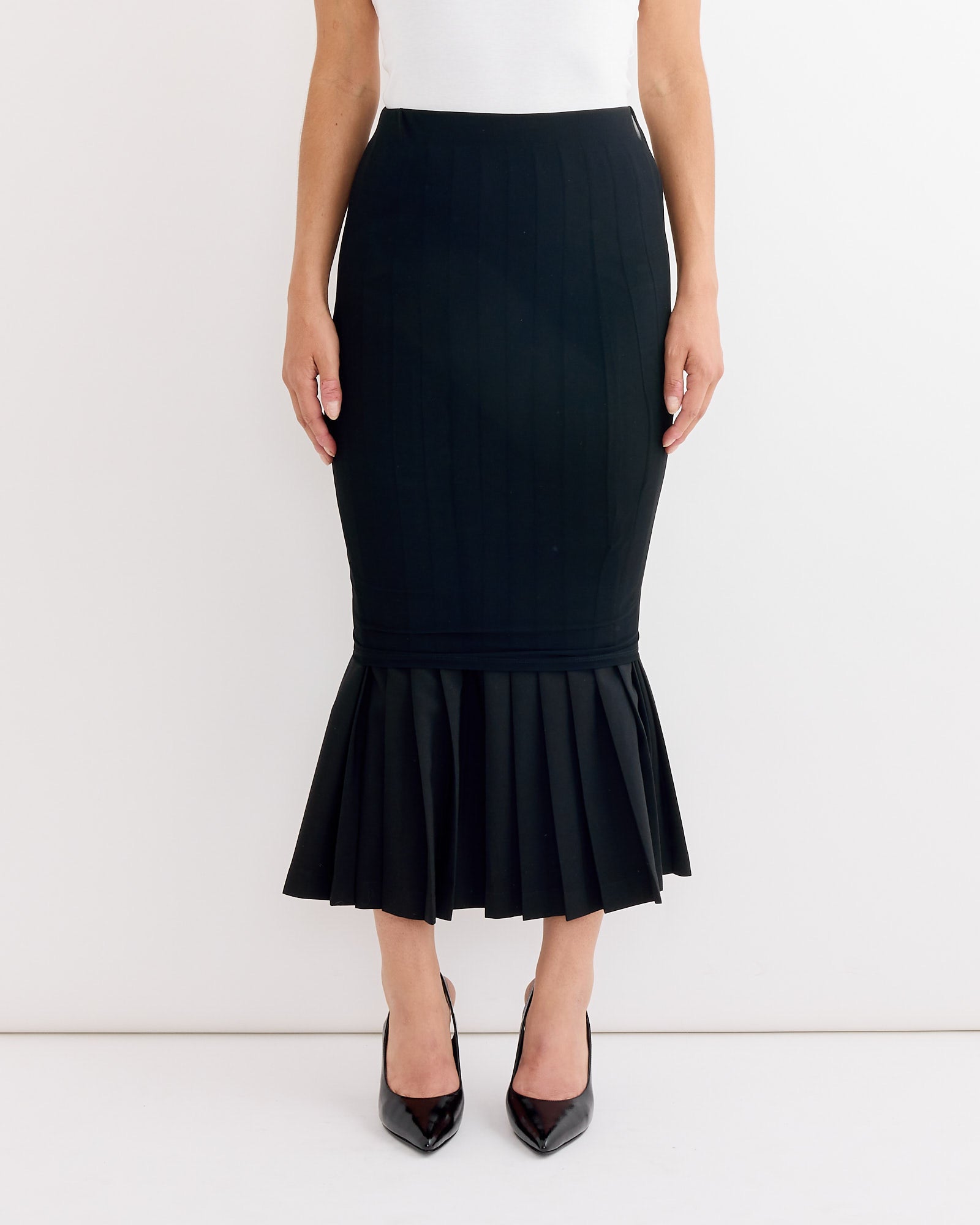 A woman stands against a white background wearing MM6 Maison Margiela’s Long Skirt in Black with a white top and black pointed-toe heels. The skirt features a fitted silhouette and pleated flared hem.