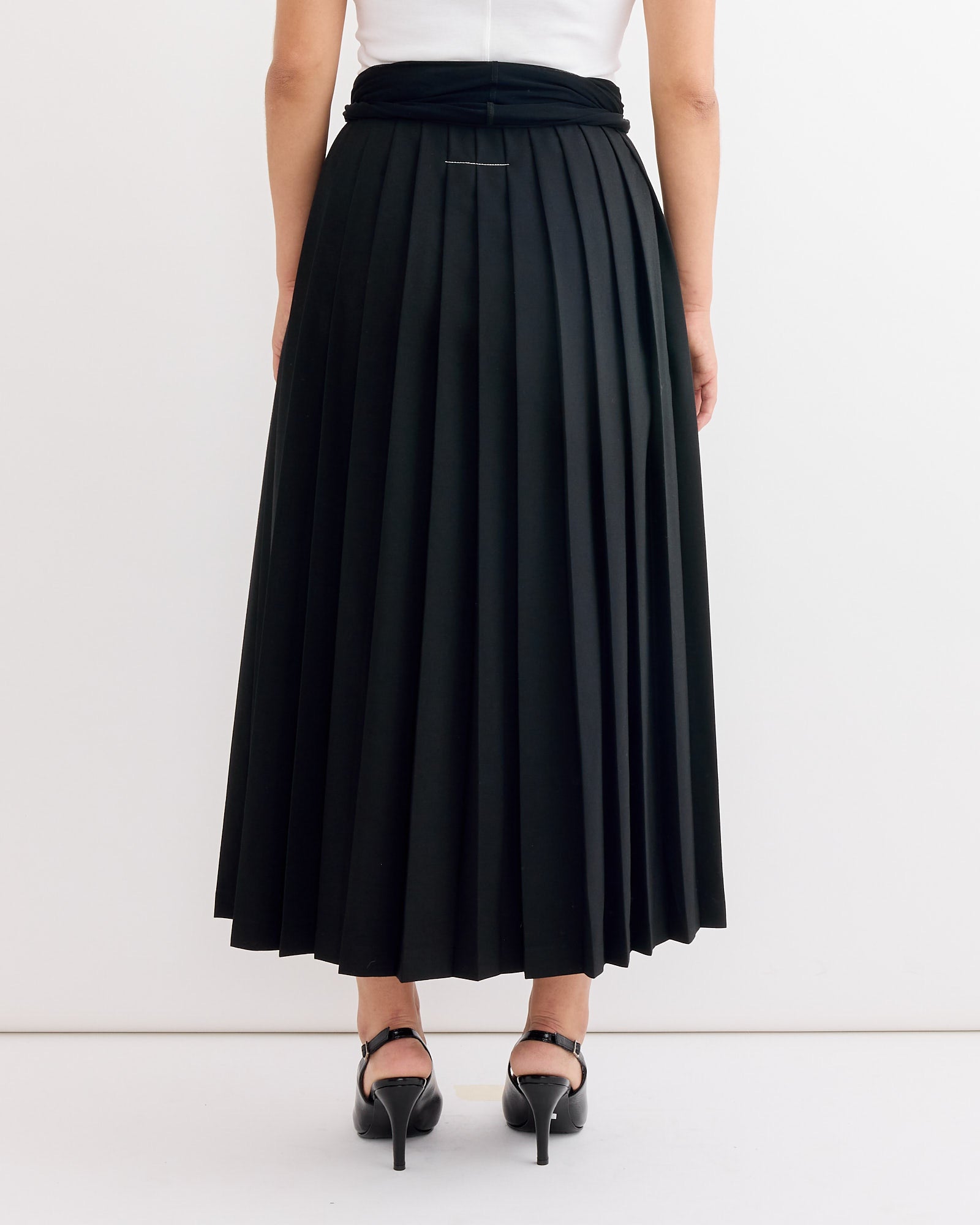 A person is shown from the back wearing a white top, MM6 Maison Margiela's Long Skirt IN Black, and black high-heeled shoes, standing against a plain white wall.
