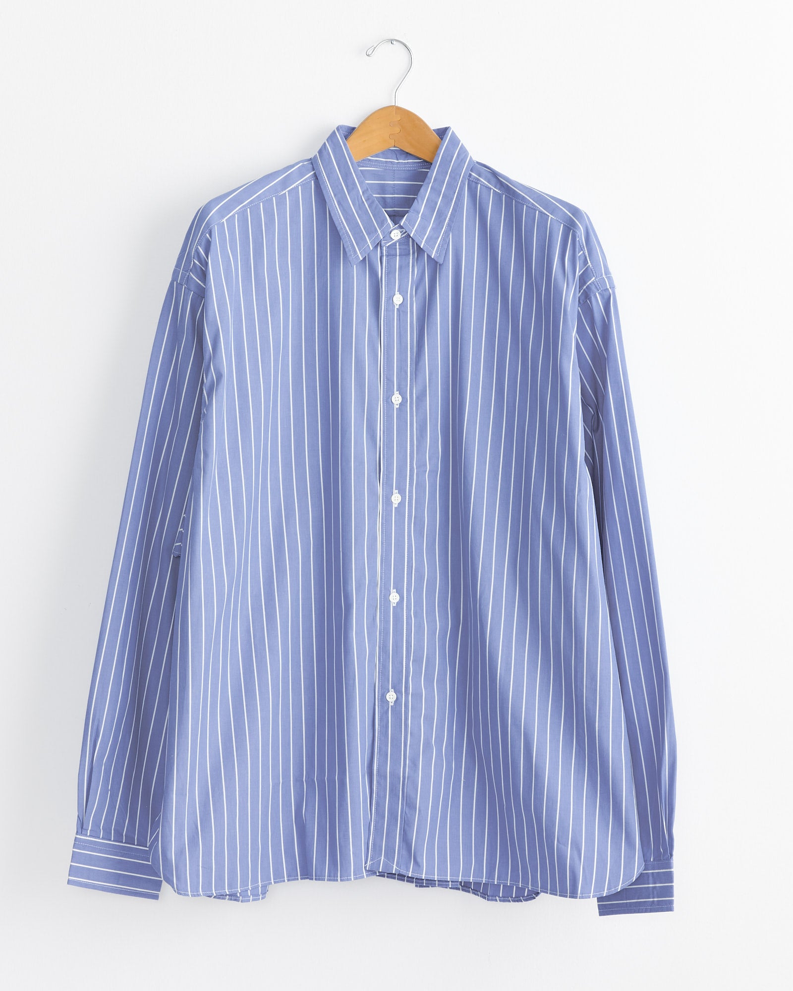 The Sillage Regular Wide High Count Shirt in White Stripe hangs on a wooden hanger against a white wall, showcasing the brand's classic style.