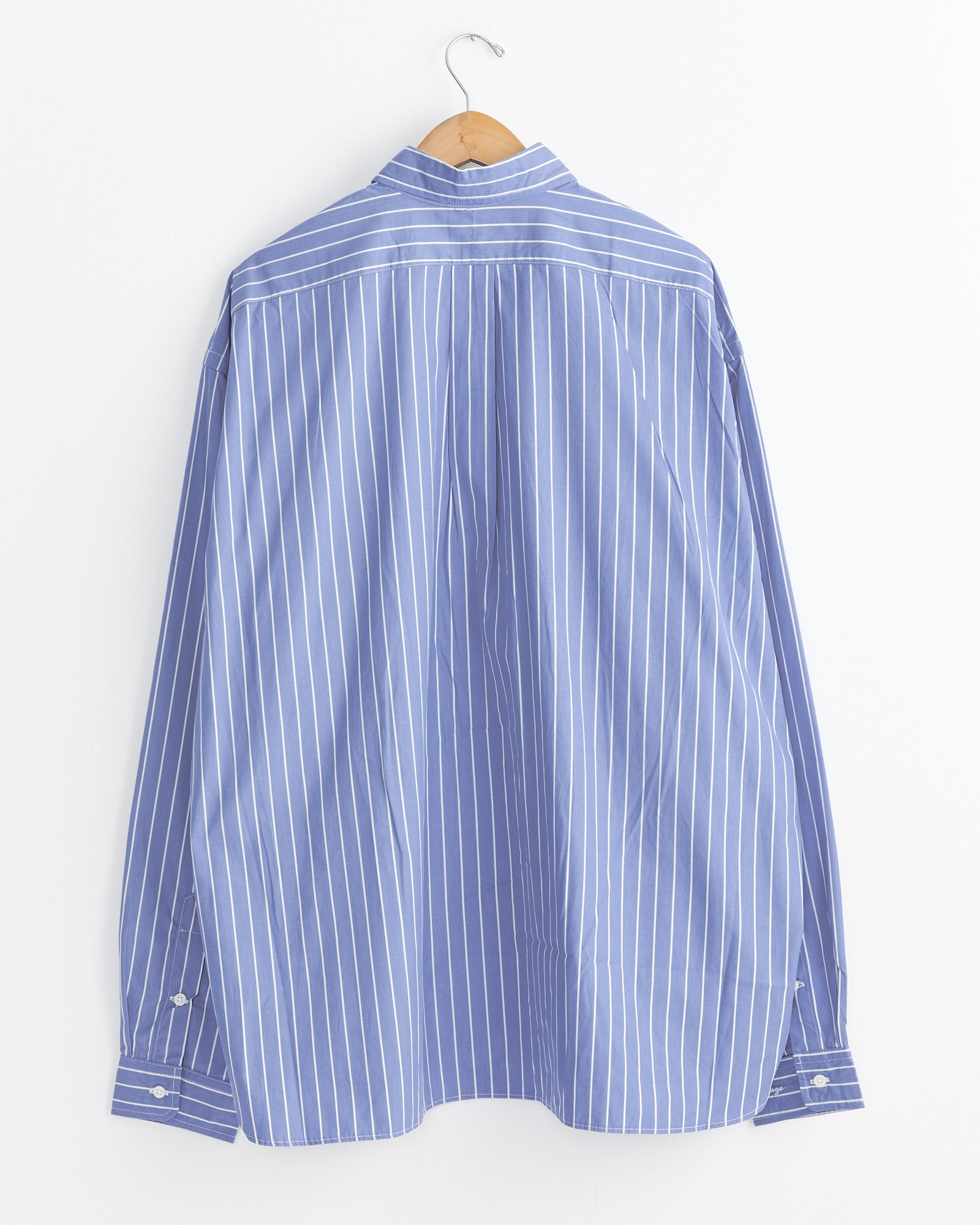 The Sillage Regular Wide High Count Shirt in White Stripe is shown from the back, hanging on a wooden hanger against a plain white wall.