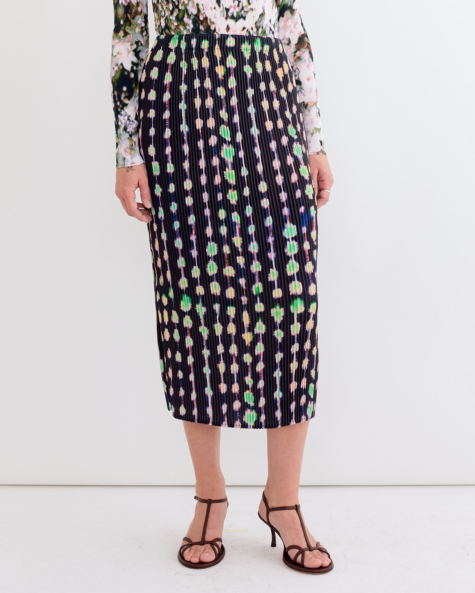 A person wears the Julia Heuer Ekke Skirt in Dax, featuring colorful geometric dots and black stripes in signature Kristall pleating, paired with a floral long-sleeve top and black strappy sandals against a plain white background.