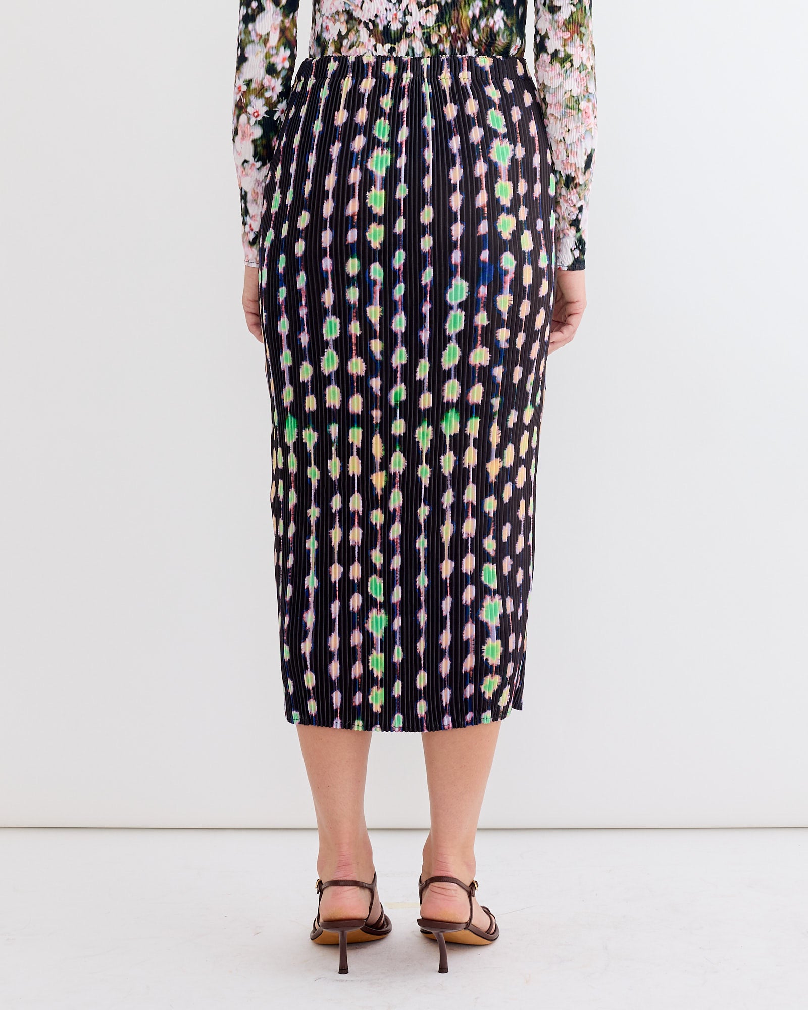 Someone wearing the Julia Heuer Ekke Skirt in Dax, featuring colorful floral and polka dot patterns, paired with a floral long-sleeve top and brown heeled sandals, stands with their back to the camera against a plain white background.