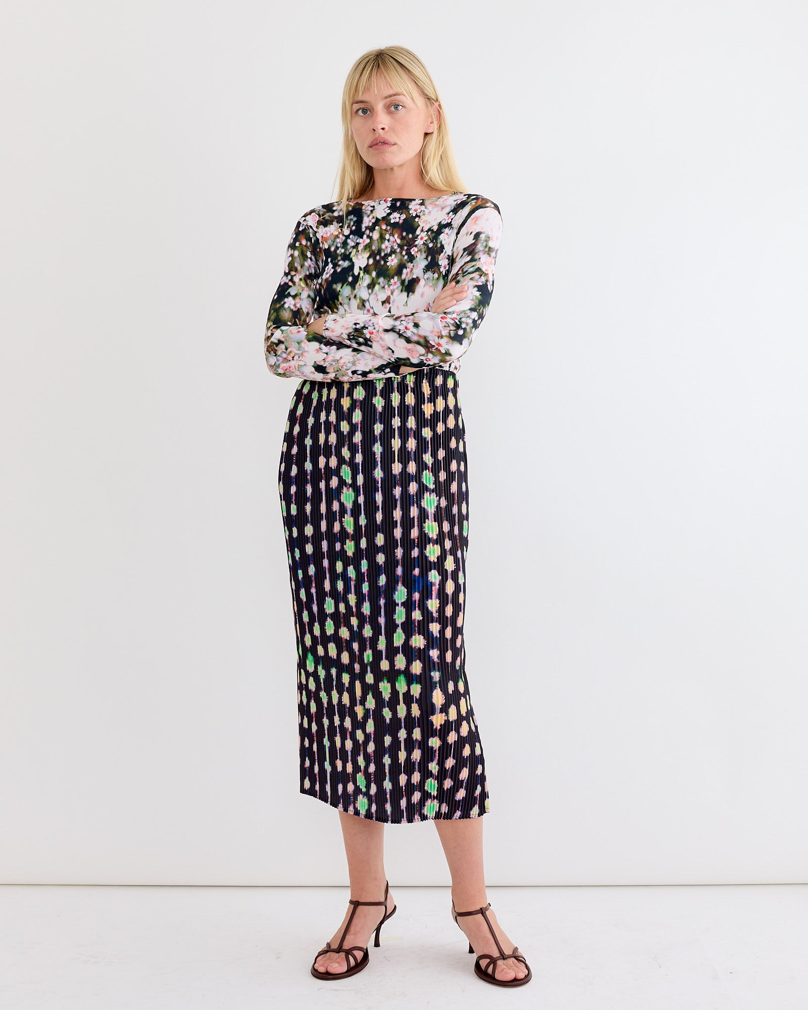 A woman stands against a plain white wall, arms crossed. She wears a long-sleeved floral top and the Julia Heuer Ekke Skirt in Dax with Kristall pleating in a pencil silhouette, paired with black strappy sandals.