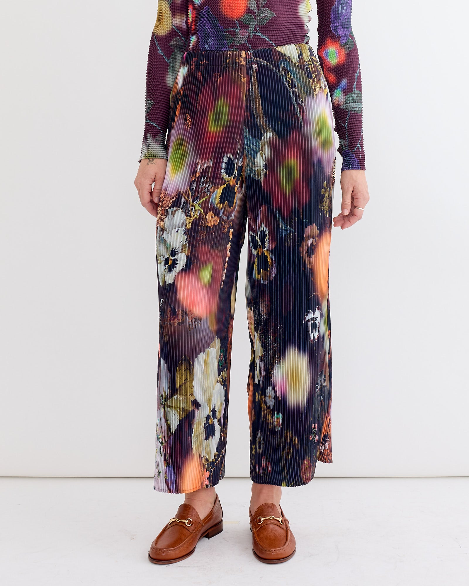 A person wears Julia Heuer’s Jack Trouser in Nola—wide-leg, hand-pleated trousers with a colorful floral pattern—paired with brown loafers and a matching vibrant top, set against a plain white background.