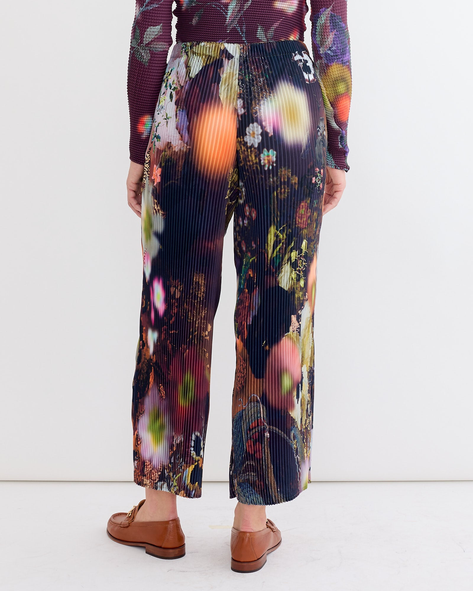 A person faces away, wearing the Julia Heuer Jack Trouser in Nola—a hand-pleated pant with a colorful abstract floral pattern—paired with brown flats and a long-sleeved patterned top, set against a plain white background.