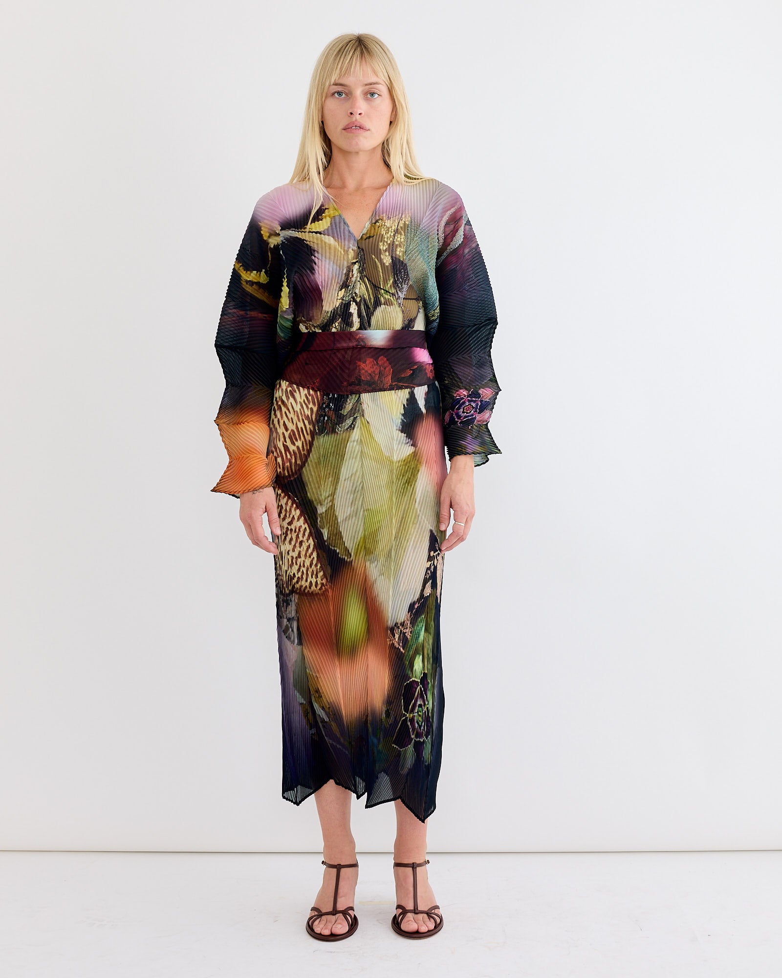 A woman with straight blonde hair and brown sandals stands against a white wall, wearing the Julia Heuer Sue Kaftan in Nola—a vibrant, hand-pleated dress featuring abstract floral and nature-inspired patterns and a reversible V-neckline.