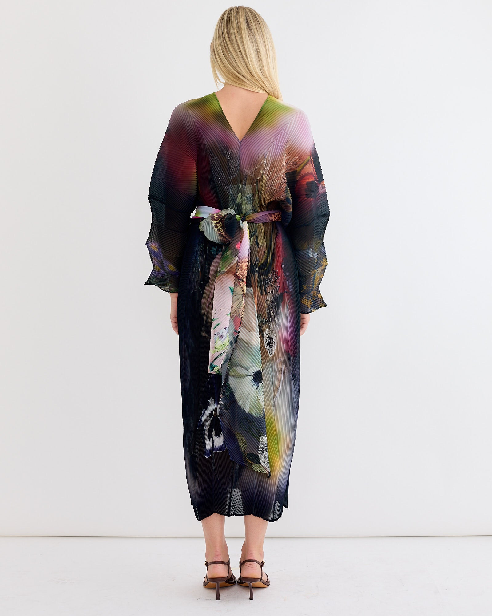 A blonde woman faces away, wearing the Julia Heuer Sue Kaftan in Nola—a long, flowy, multicolored dress with a reversible V-neck and floral sash—paired with brown strappy heels, set against a plain white background.