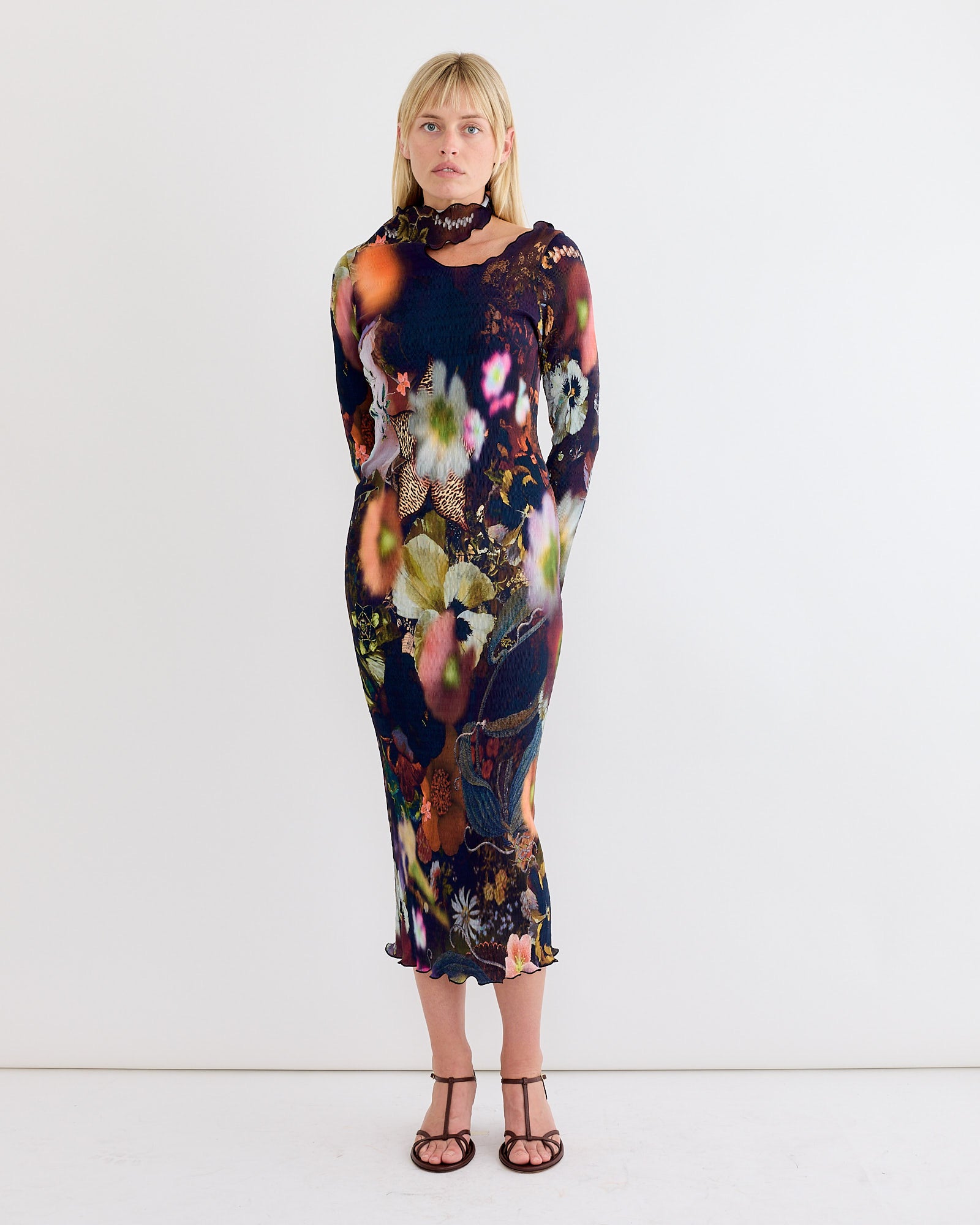 A woman stands against a white wall wearing the Julia Heuer Ophelia Dress in Nola, a hand-pleated, long-sleeve midi dress in multicolored floral recycled polyester crepe, paired with open-toed sandals and straight blonde hair.