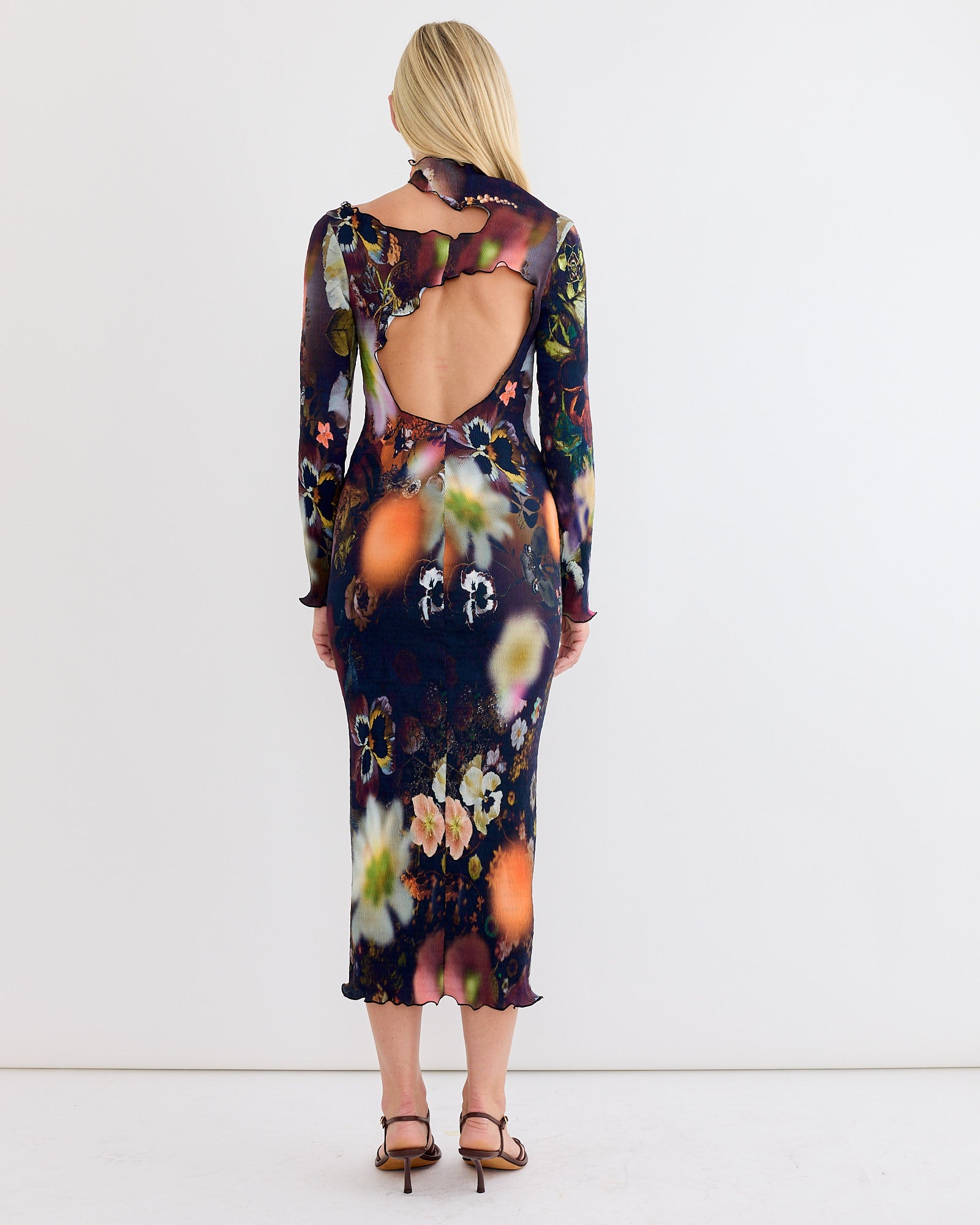 A woman with long blonde hair stands before a white wall in strappy heeled sandals, wearing the Julia Heuer Ophelia Dress in Nola—a colorful, hand-pleated, open-back, long-sleeve midi dress.