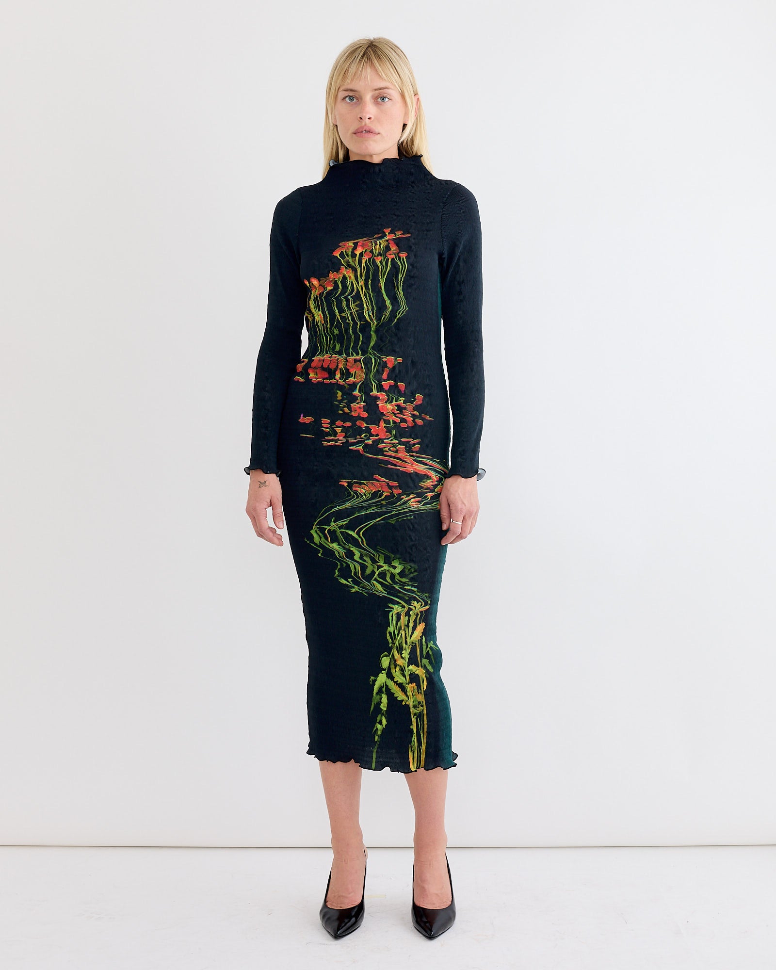 A woman with light, straight shoulder-length hair and a neutral expression stands against a white wall, wearing the Julia Heuer Rupert Dress in Patsy—long-sleeved, recycled polyester georgette with a colorful abstract vertical pattern—and black heels.