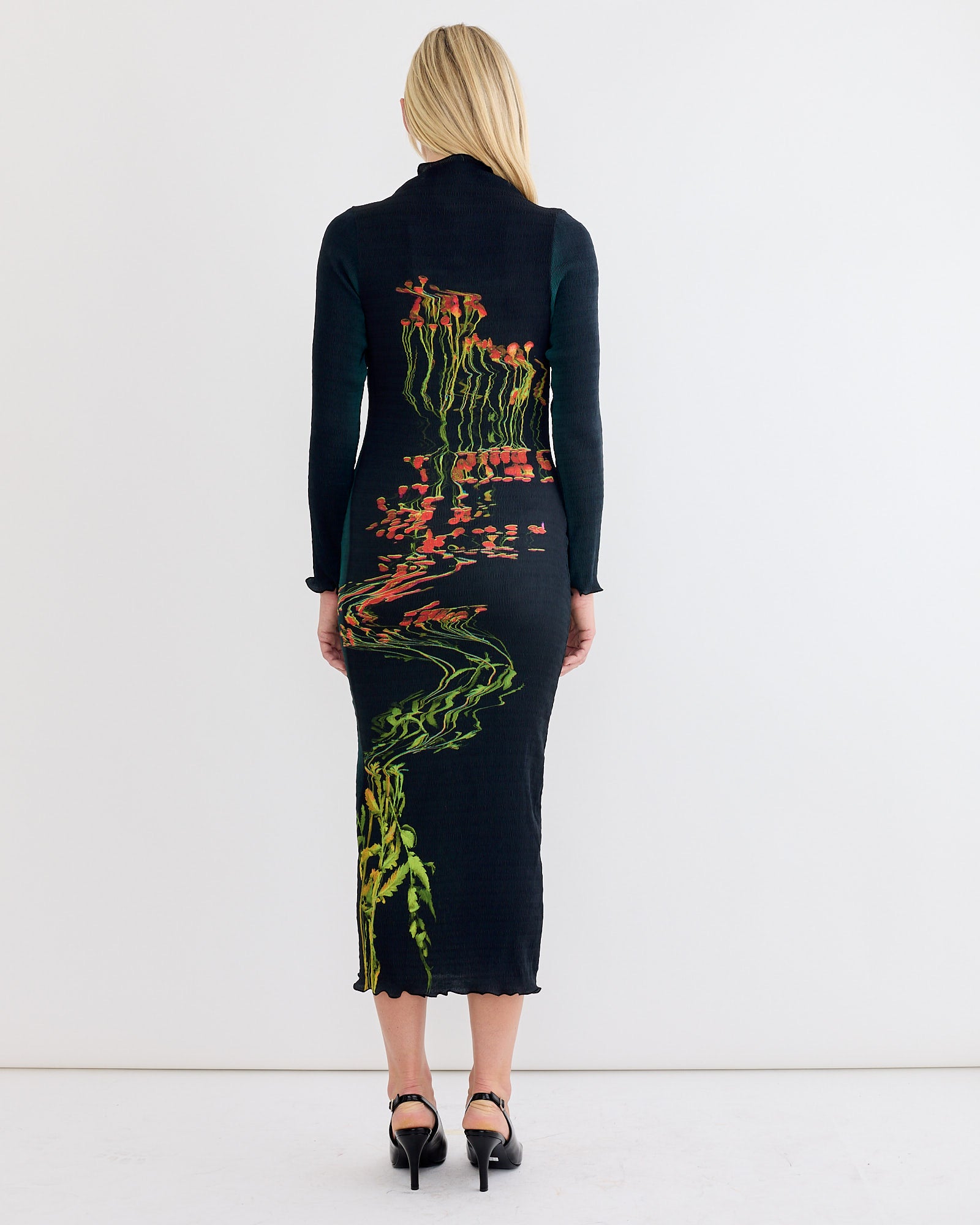 A woman with long blonde hair, wearing the Julia Heuer Rupert Dress in Patsy—black with colorful florals and green stems on the back—stands facing away in a minimalist white room. The dress features elegant smock pleat detailing.