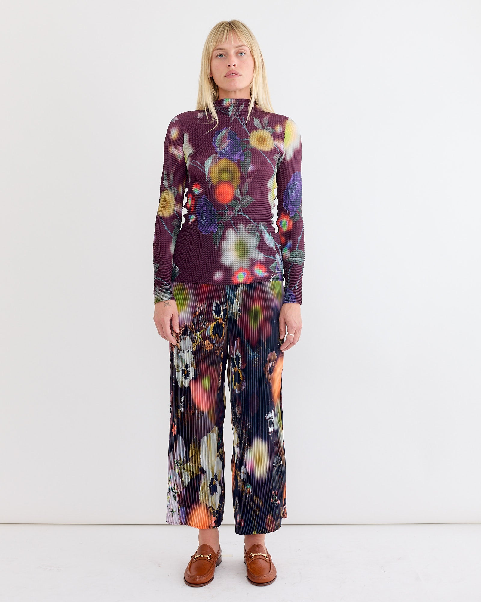 A blonde woman wears the hand-pleated Susi Top in Edward and matching wide-leg pants by Julia Heuer, both featuring a colorful abstract floral print, paired with brown loafers, against a plain white background.