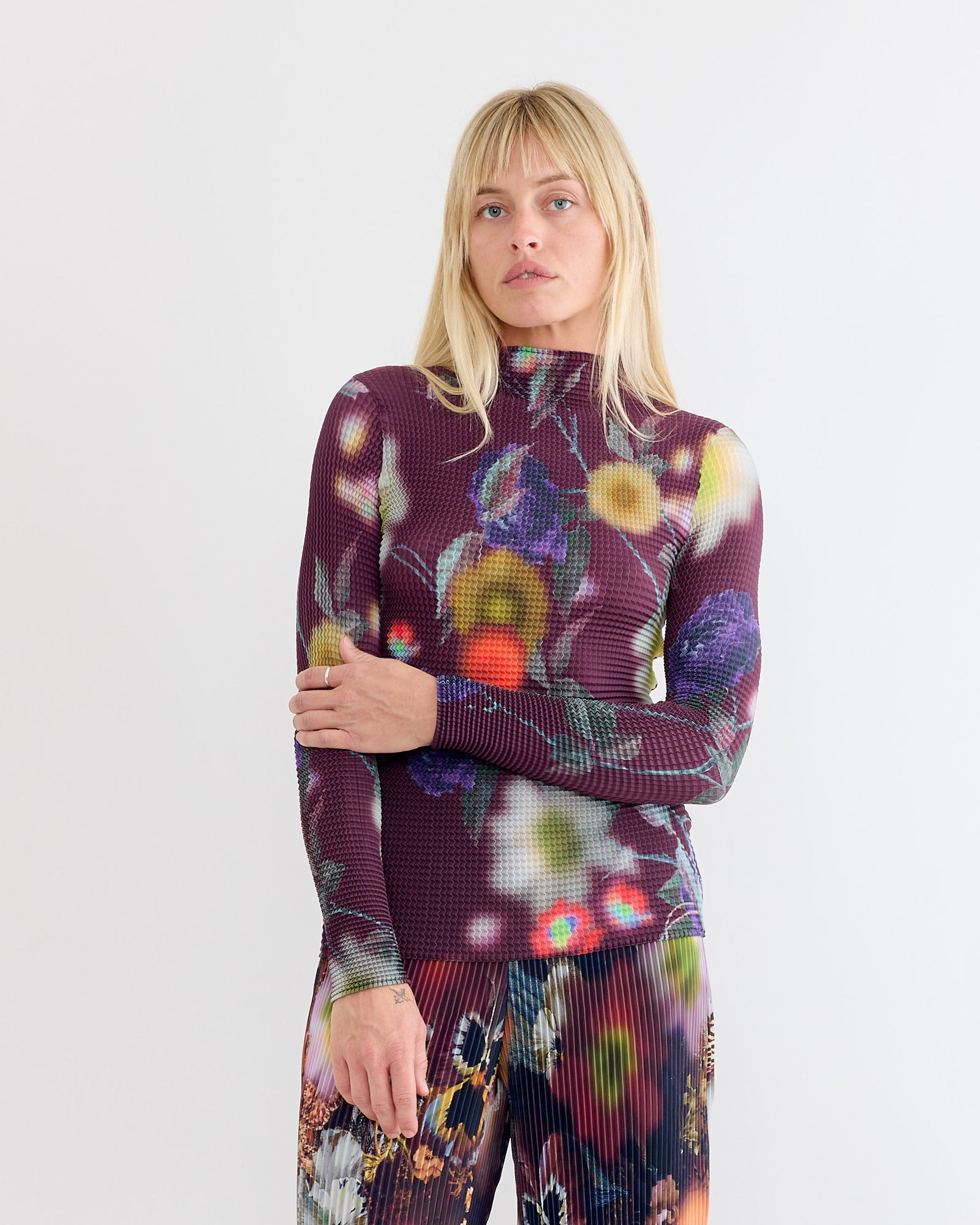 A blonde-haired woman with bangs wears the Julia Heuer Susi Top in Edward, a fitted, long-sleeve, high-neck piece with a colorful abstract floral pattern and hand-pleated texture, styled with matching pants against a plain white backdrop.
