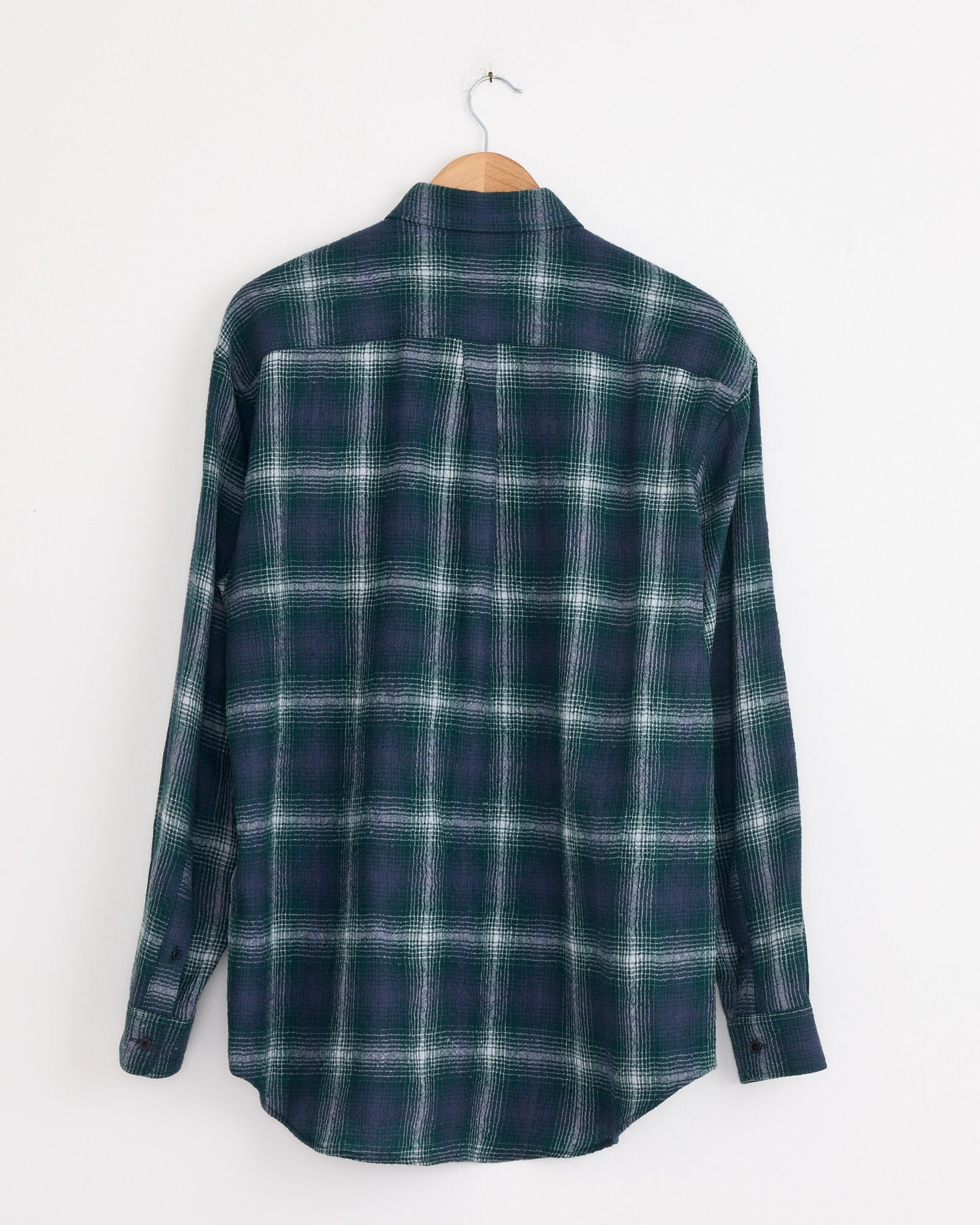 An Auralee Airy Wool Check Shirt in Forest Green/Navy hangs on a wooden hanger against a white wall, shown from the back. Made in Japan from soft, airy wool for comfort and style.