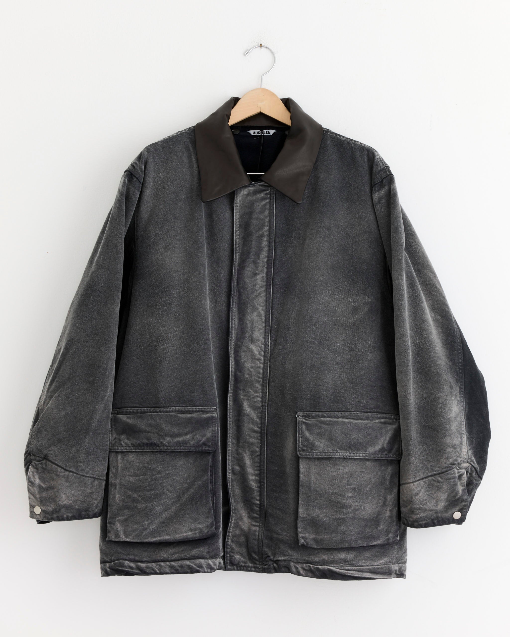 The Auralee Dyed Duck Blouson in Faded Black, featuring a dark collar, large front pockets, and snap-button cuffs, crafted from organic cotton, hangs on a wooden hanger against a white background.