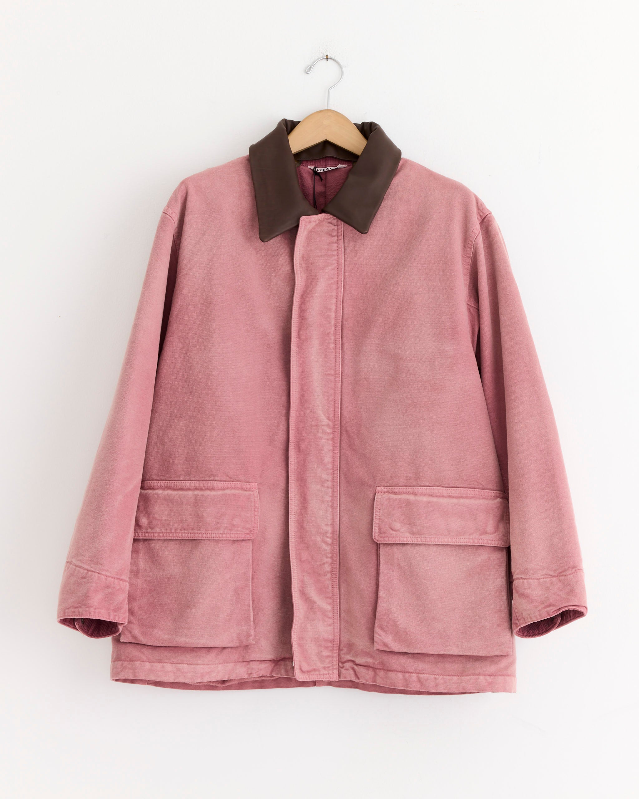 The Auralee Dyed Duck Blouson in Faded Burgundy, featuring a brown collar and two large front pockets, hangs on a wooden hanger against a white background.