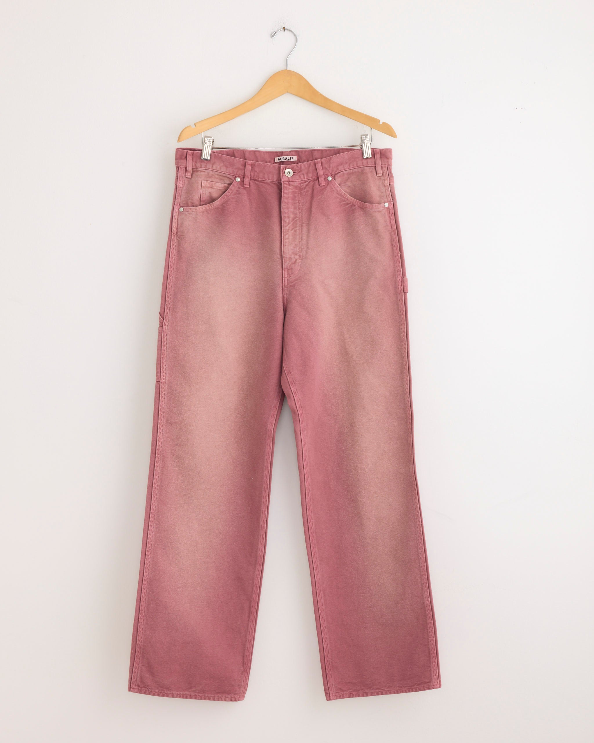 A pair of Auralee Dyed Duck Painter Pant in Faded in Burgundy, crafted from organic cotton duck, hangs on a wooden hanger against a plain white wall.