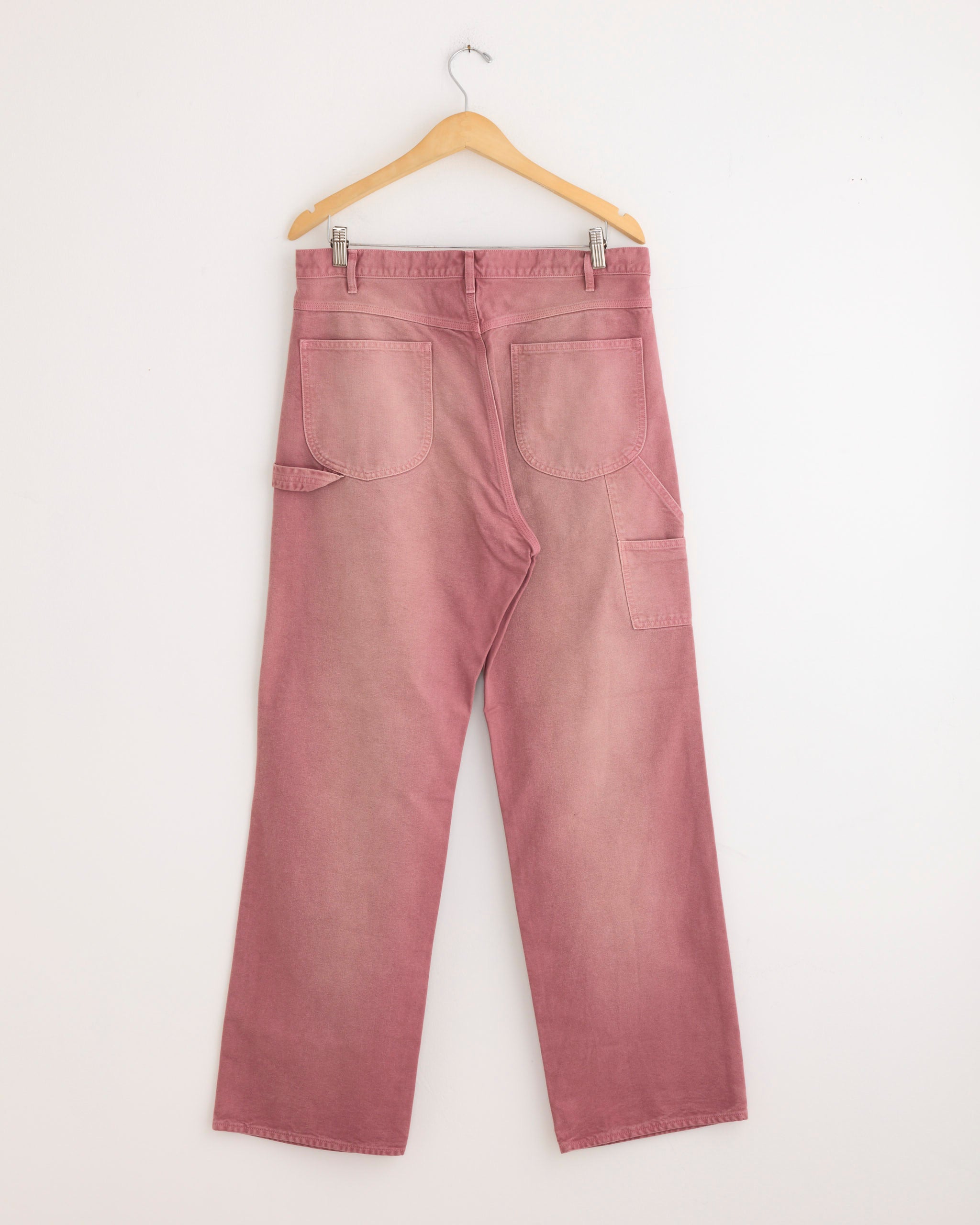 A pair of Auralee Dyed Duck Painter Pants in Faded Burgundy, featuring wide legs, large back pockets, and a small utility pocket in organic cotton duck, shown on a wooden hanger against a white wall.