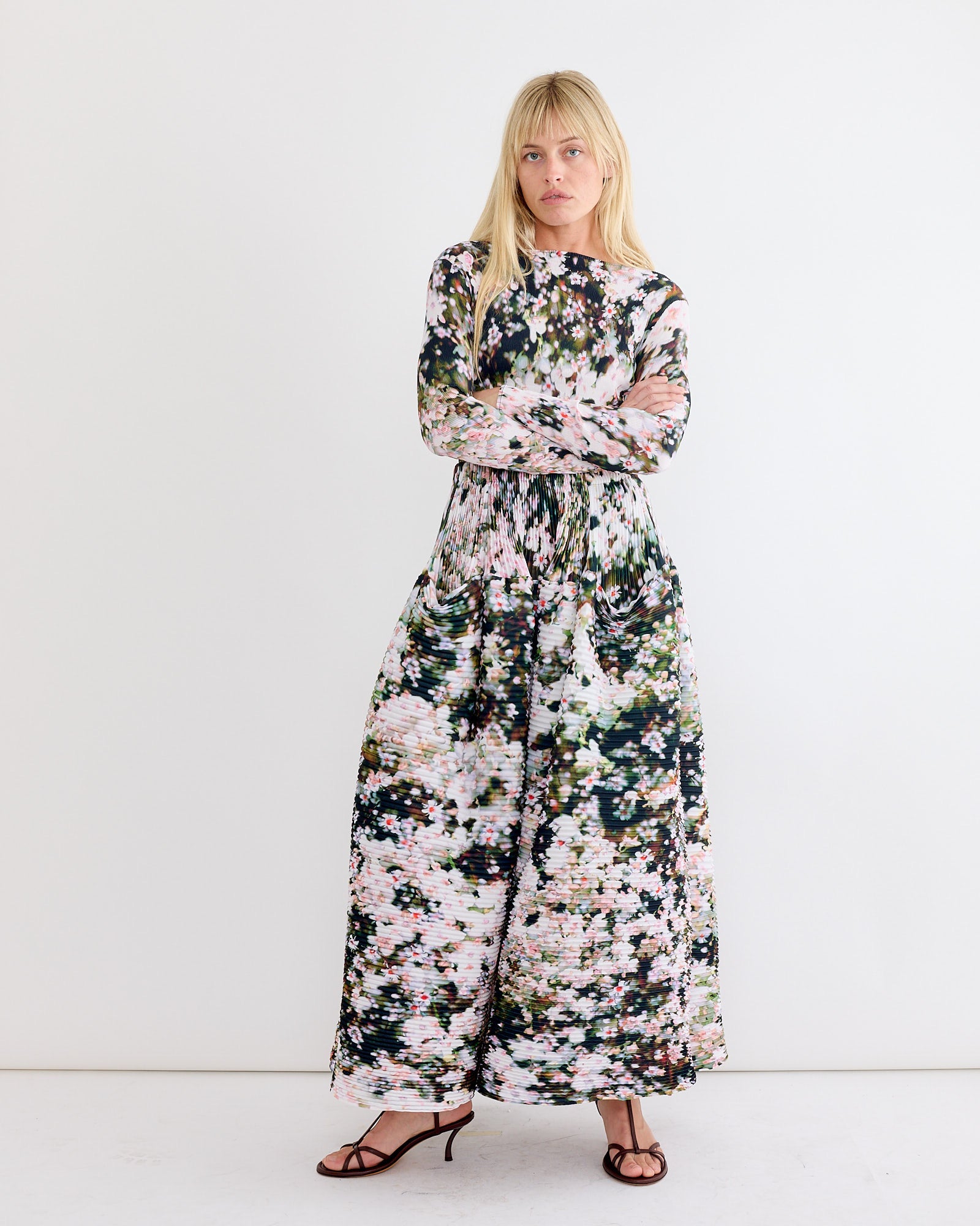 A person with long blonde hair stands against a white wall, wearing the Julia Heuer Kenza Skirt in Holle— a long, flowy, hand-pleated design featuring abstract floral patterns in pink, green, and black, paired with dark strappy heels.