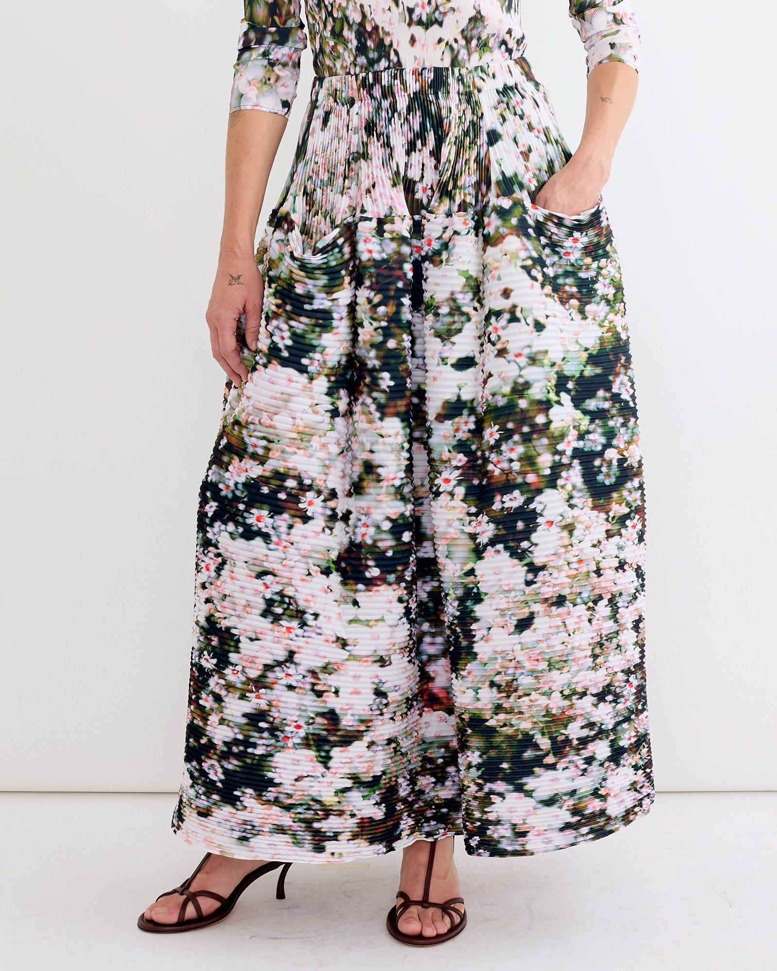 A person showcases the lower half in Julia Heuer's Kenza Skirt in Holle—a long, hand-pleated piece with green, white, and pink abstract floral patterns—standing against a simple light background.