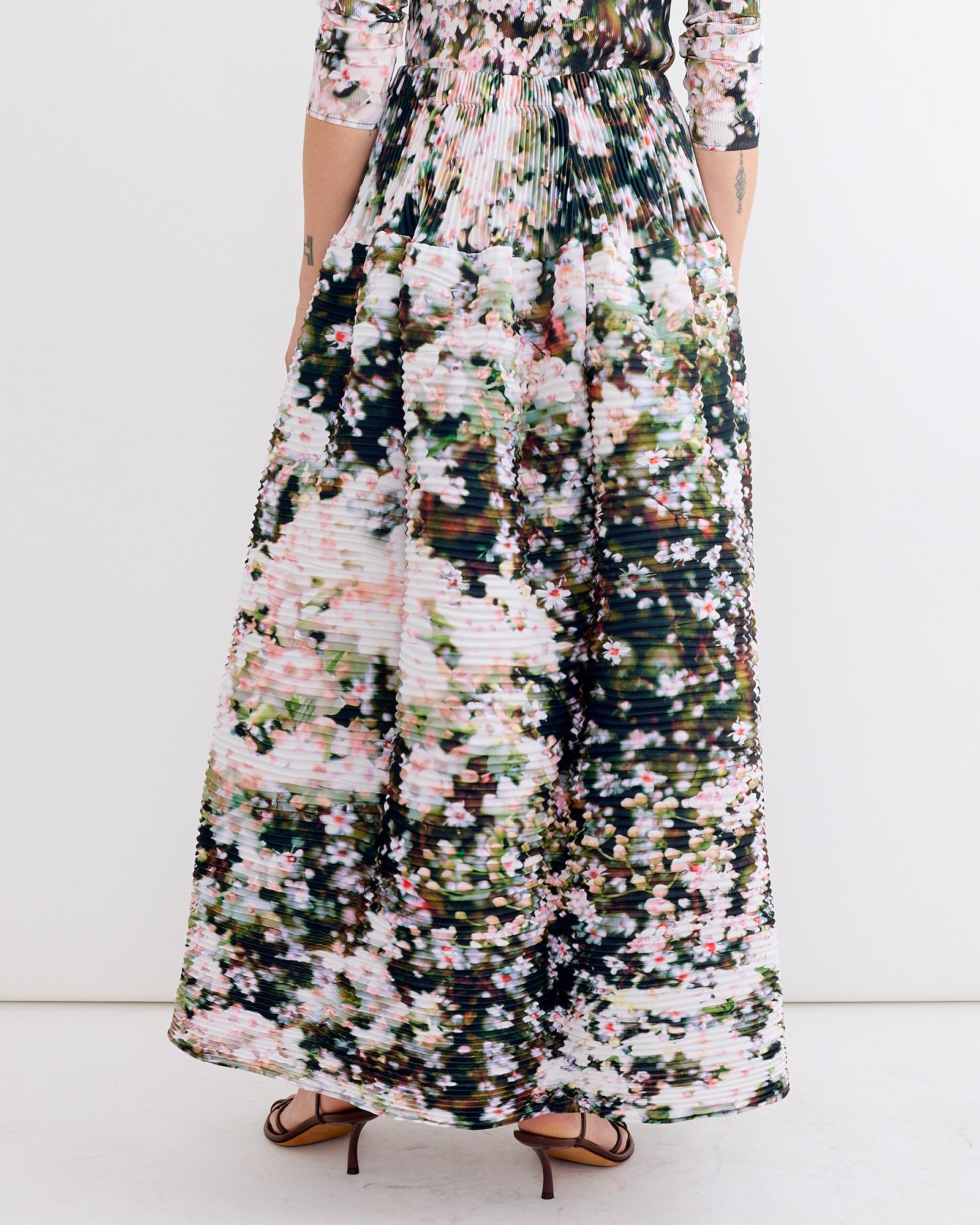 A person models the Julia Heuer Kenza Skirt in Holle, featuring hand-pleated pink, white, and green florals, standing with their back to the camera in heeled sandals against a plain white backdrop.