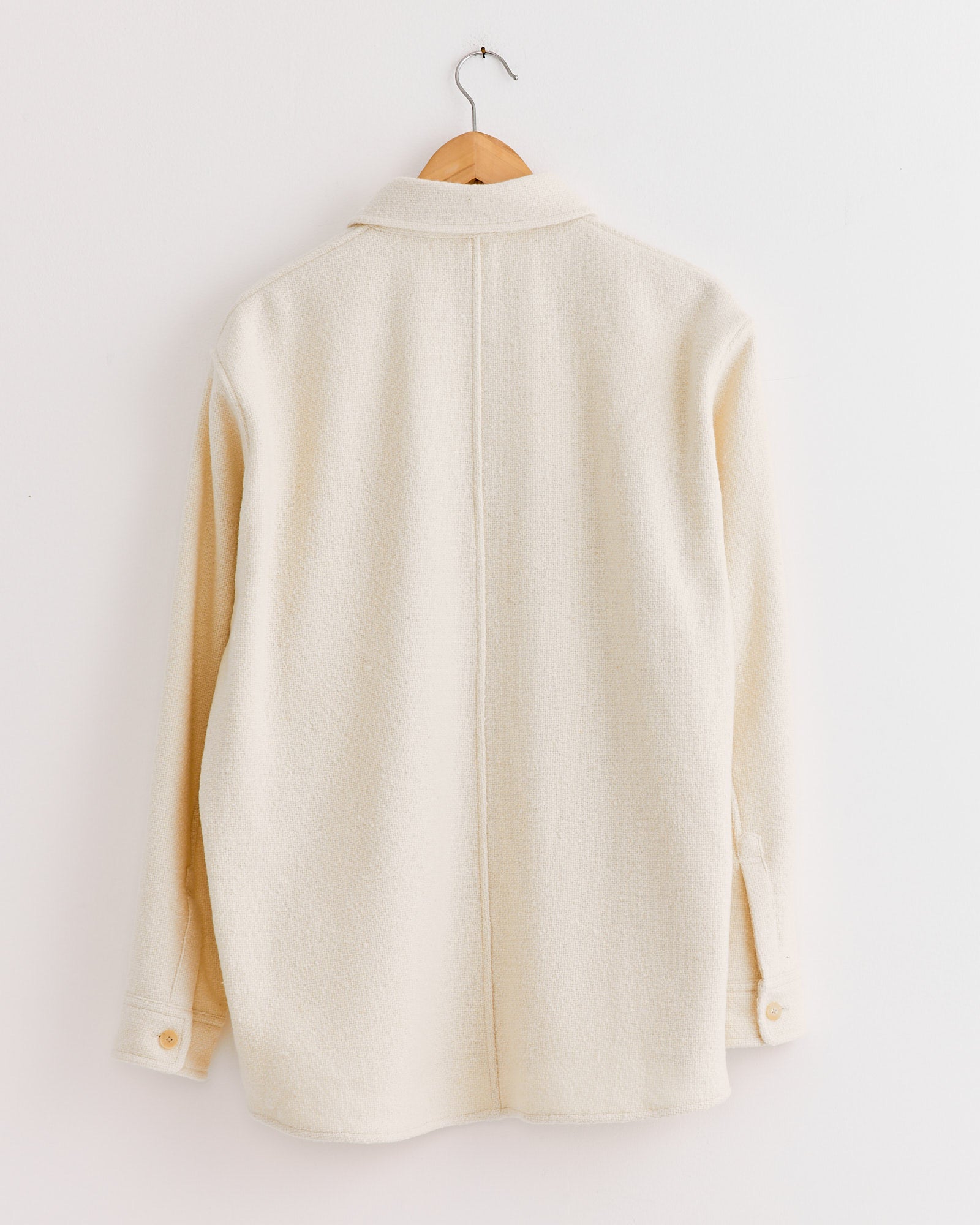 An Auralee Homespun Tweed Shirt Blouson in Ivory White hangs on a wooden hanger against a white wall, shown from the back. Made in Japan with Super 120’s wool, this soft, textured shirt features cuff buttons.