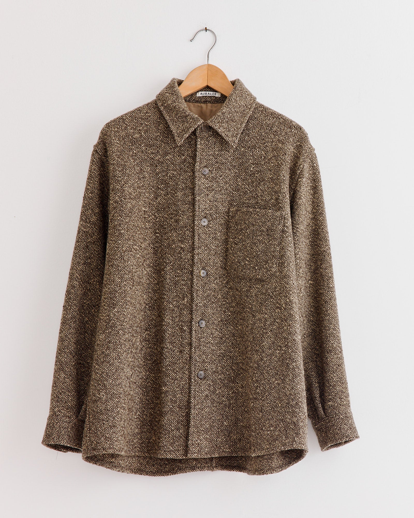 The Auralee Homespun Tweed Shirt Blouson in Brown, crafted in Japan from Super 120’s wool, features a textured finish, button-up front, collar, and chest pocket—shown on a wooden hanger against a plain white wall.