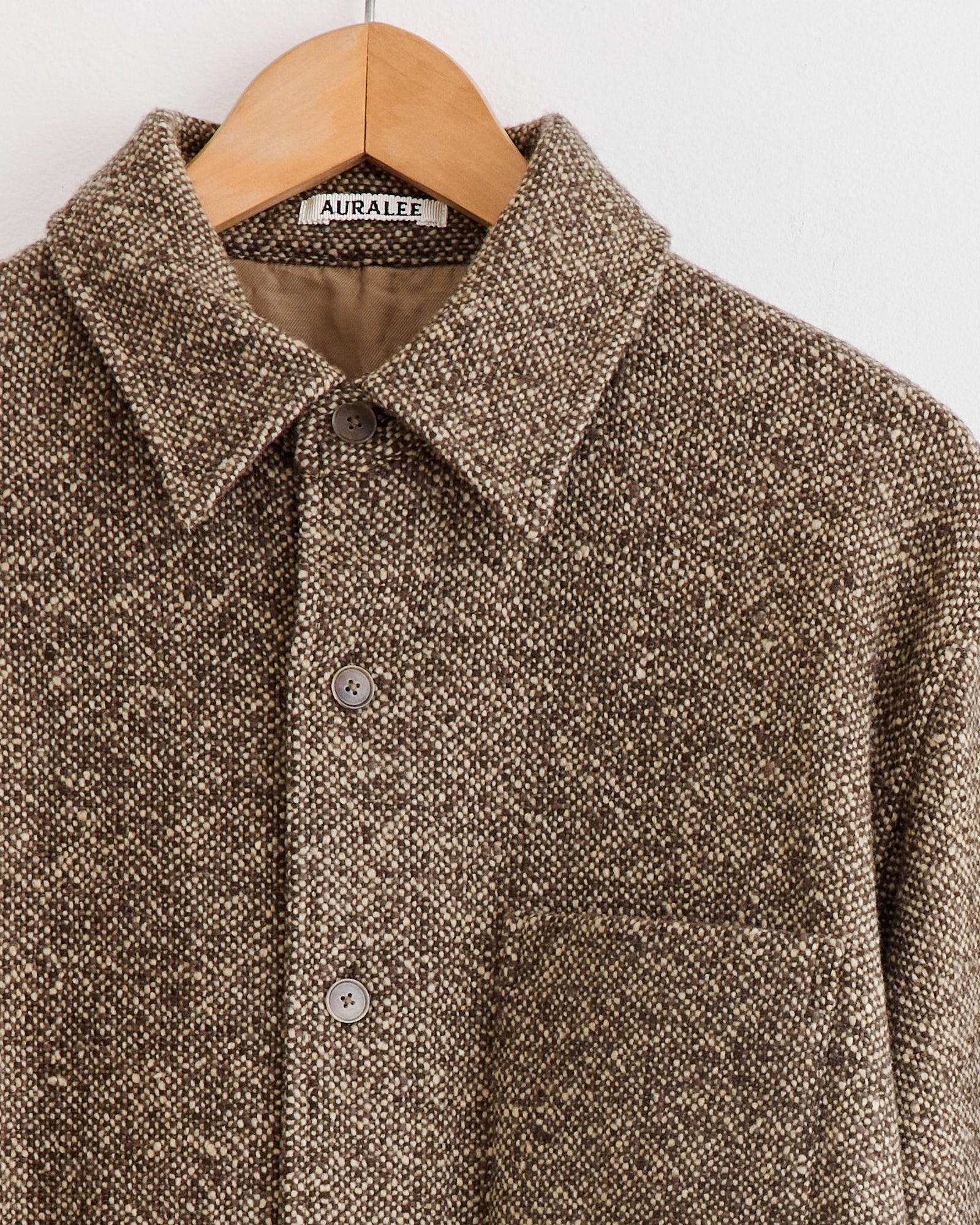 A close-up of the Auralee Homespun Tweed Shirt Blouson in Brown, featuring a collar, button-down front, and chest pocket, crafted from Super 120’s wool, hangs on a wooden hanger. The label reads Auralee. Made in Japan.