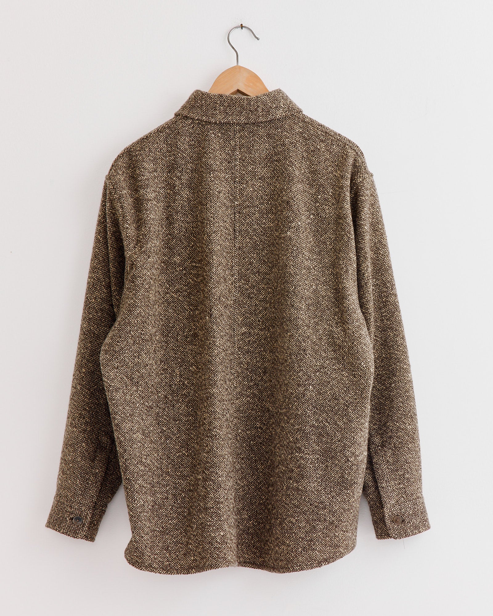 A brown Auralee Homespun Tweed Shirt Blouson hangs on a wooden hanger against a white wall, shown from the back. Made in Japan, its textured fabric adds a refined touch.