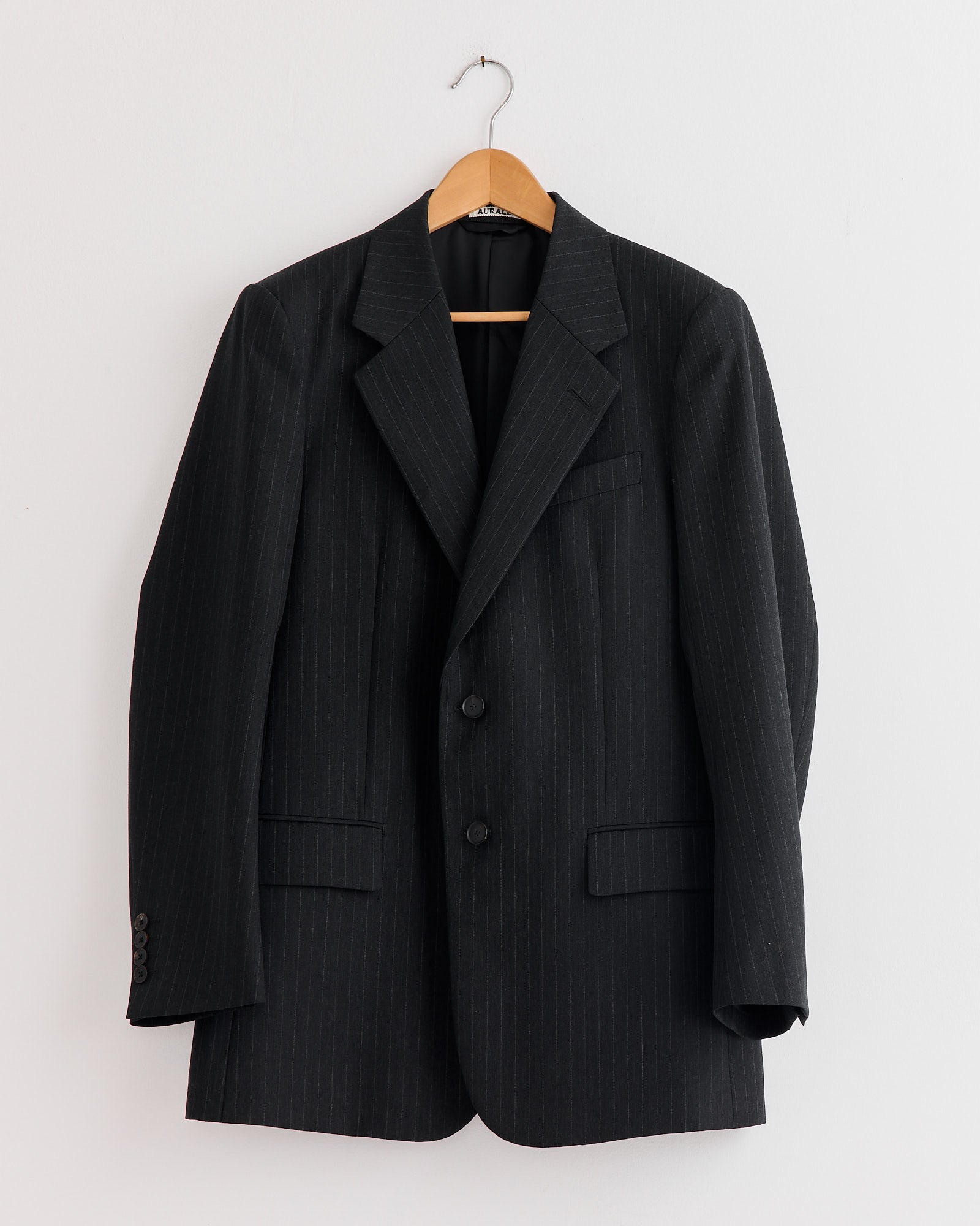 The Auralee Bluefaced Wool Jacket in Charcoal Navy Stripe, crafted in Japan from extra fine merino, features black pinstripes, notch lapels, two buttons, and front pockets. Shown on a wooden hanger against a white wall.