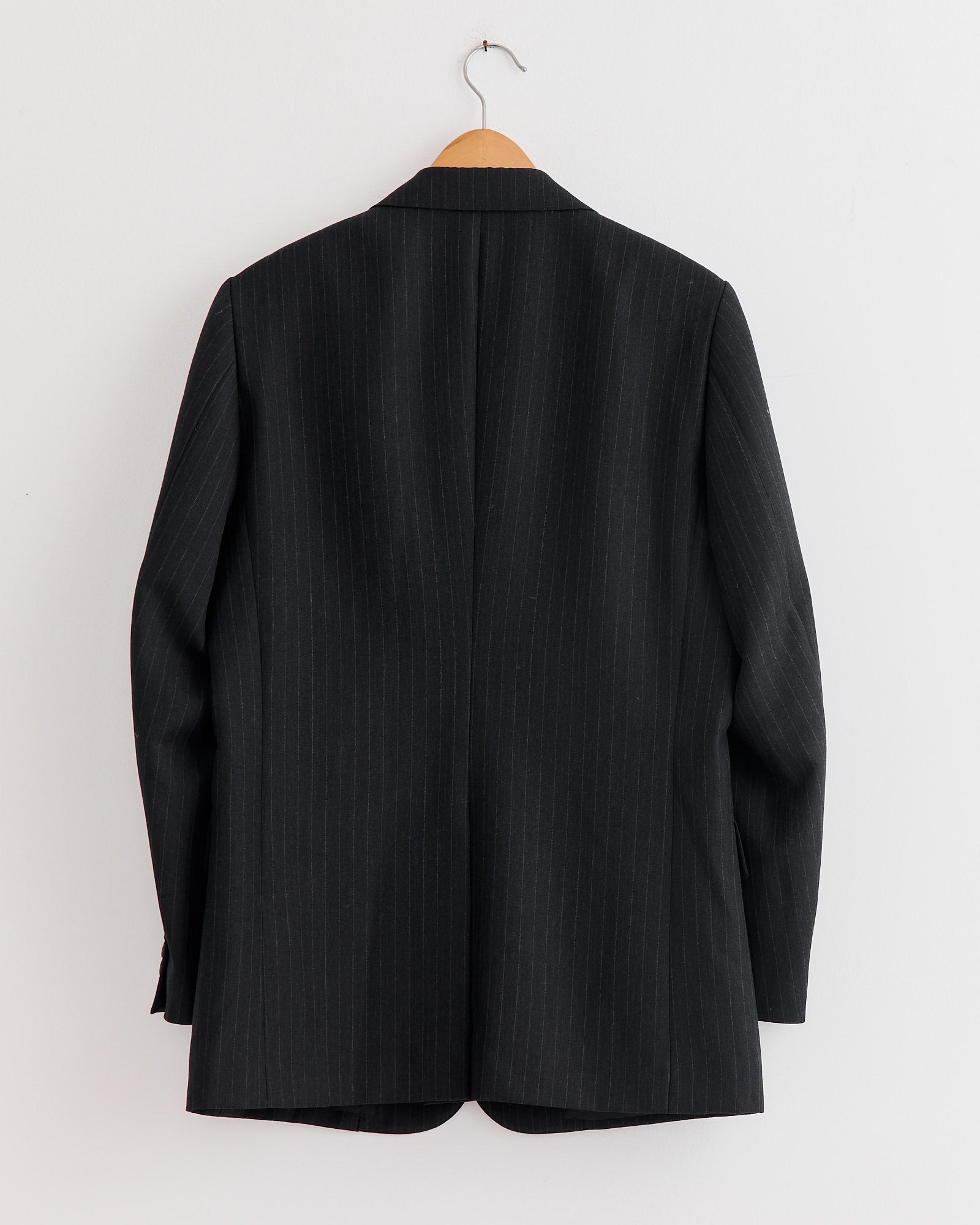 The Auralee Bluefaced Wool Jacket in Charcoal Navy Stripe, made in Japan from extra fine merino, hangs on a wooden hanger against a white wall—its back view highlighting refined craftsmanship and timeless style.