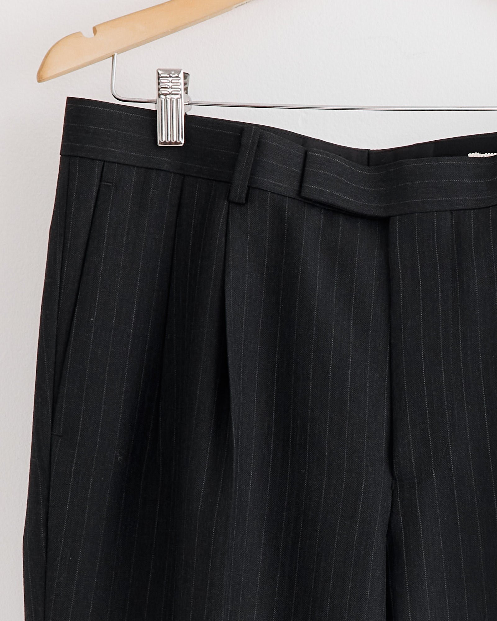 The Auralee Bluefaced Wool Two Tuck Slack in Charcoal Navy Stripe hangs on a wooden hanger against a white wall.