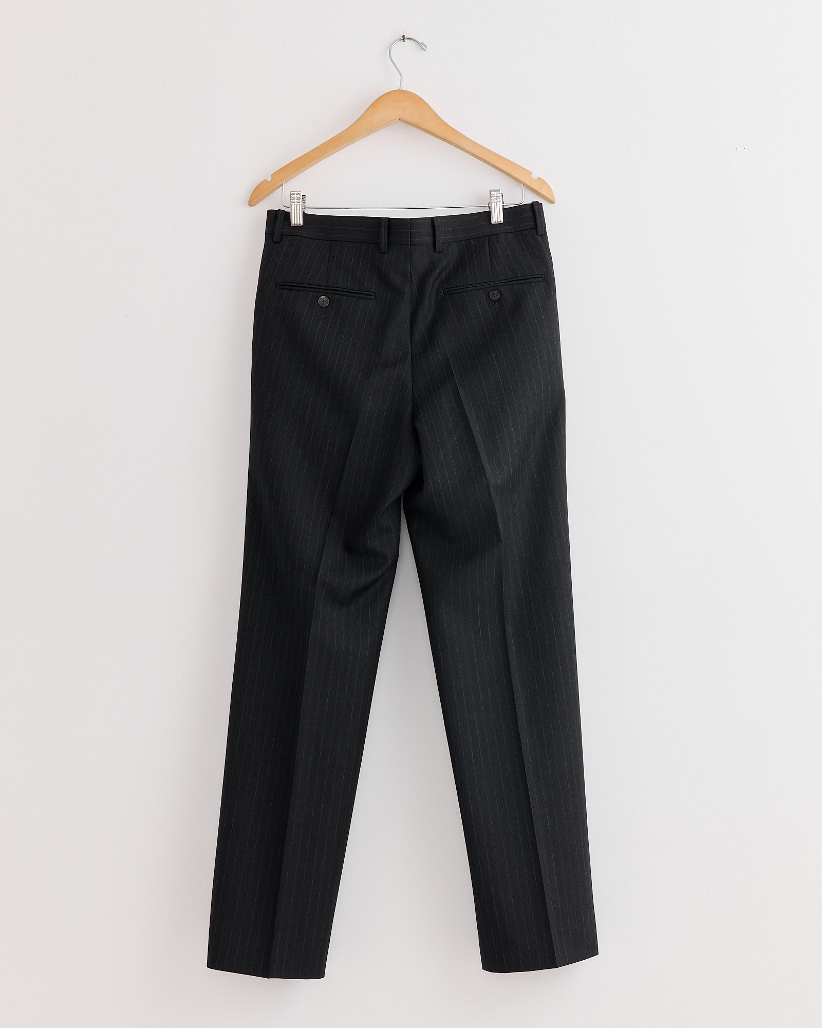A pair of Auralee Bluefaced Wool Two Tuck Slacks in Charcoal Navy Stripe hangs on a wooden hanger against a plain white wall, shown from the back.