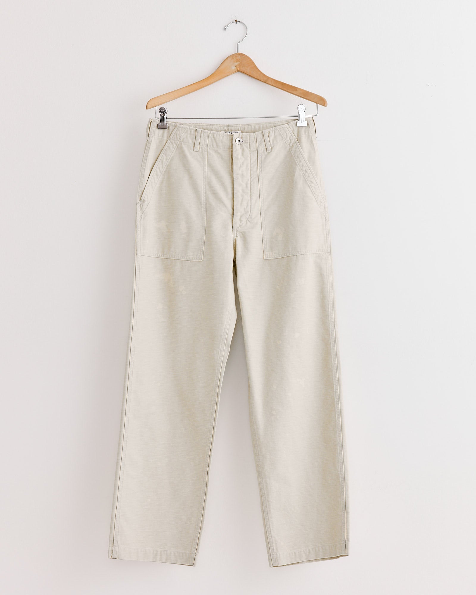 Auralee’s Washed Baggy Baker Pant in Dusty Ivory features a straight-leg silhouette, front patch pockets, button and zip closure, and belt loops, shown hanging on a wooden hanger against a plain white wall.