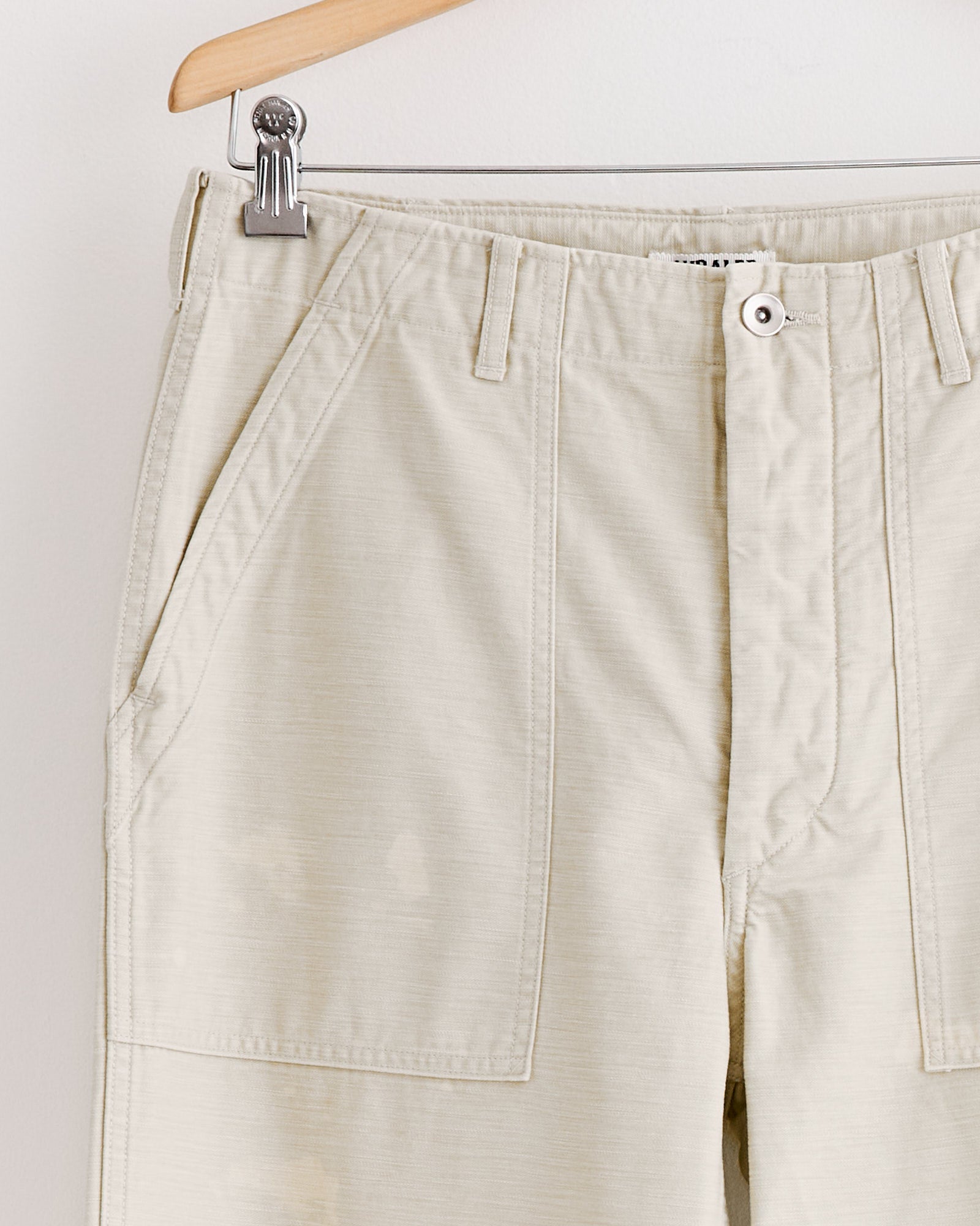 A close-up of Auralee's Washed Baggy Baker Pant in Dusty Ivory hanging on a wooden hanger with metal clips, highlighting the waistband, front pockets, and button closure against a plain white background.