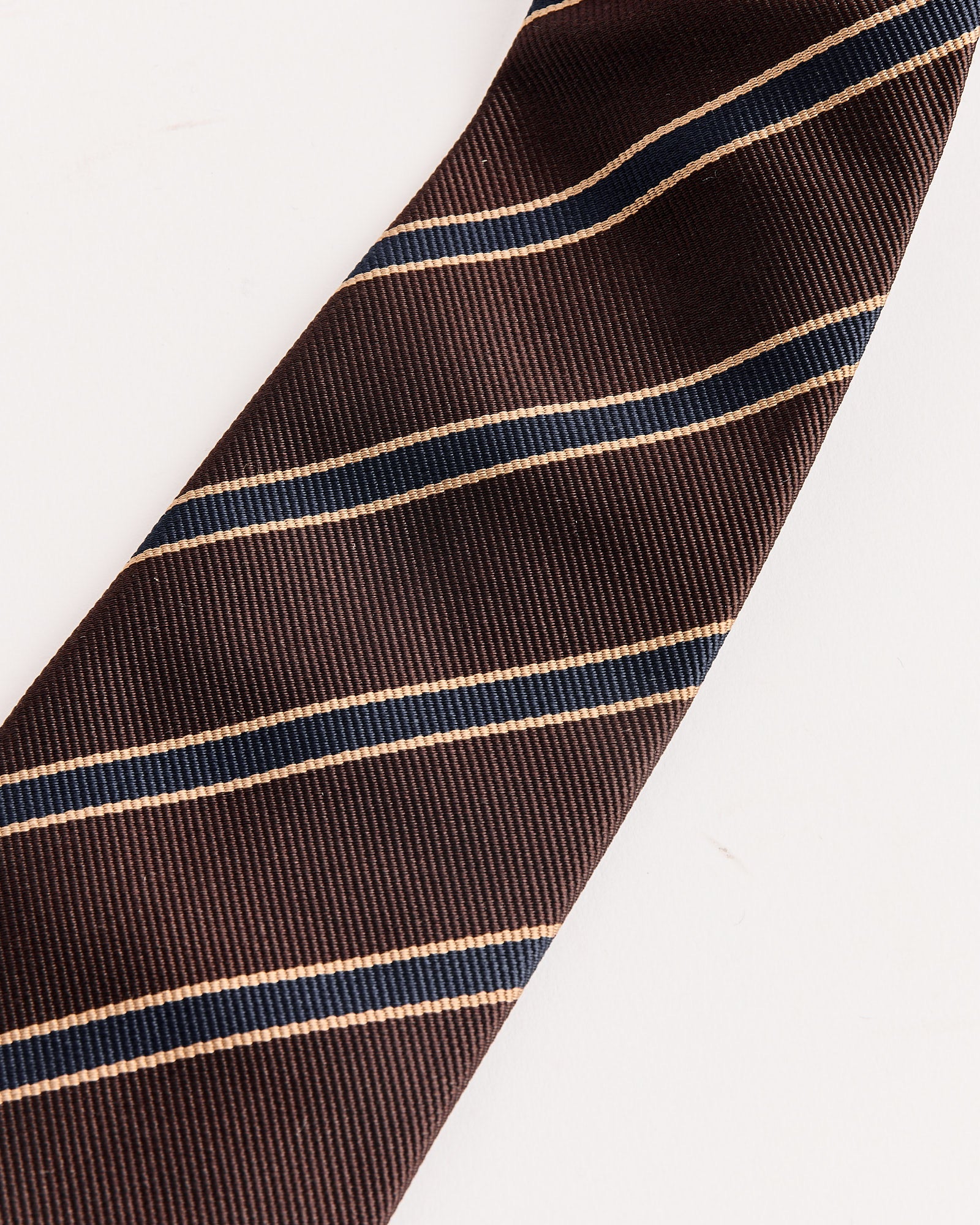 A close-up of the Auralee Silk Stripe Tie in Dark Brown, featuring diagonal beige and navy stripes, made in Japan, shown on a white background.