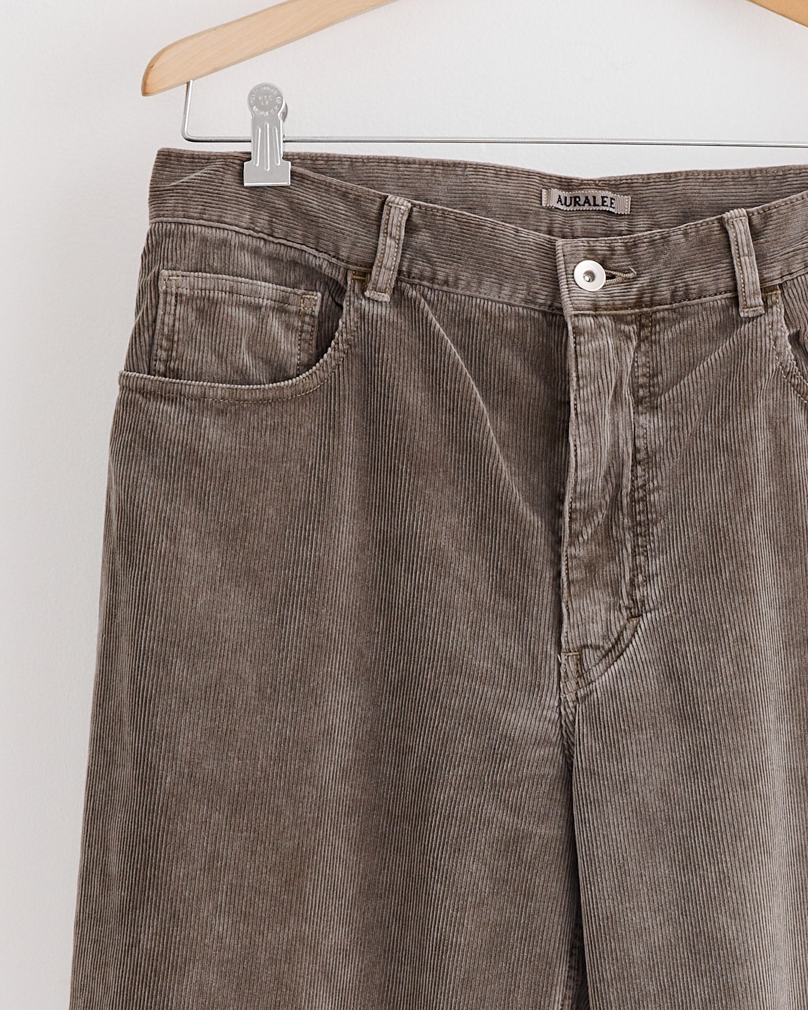 Auralee Corduroy Wide 5 Pockets Pant in Olive Brown hangs on a wooden hanger against a white wall, with the brand label visible on the inner waistband. For fit or sizing details, connect via our online chat.