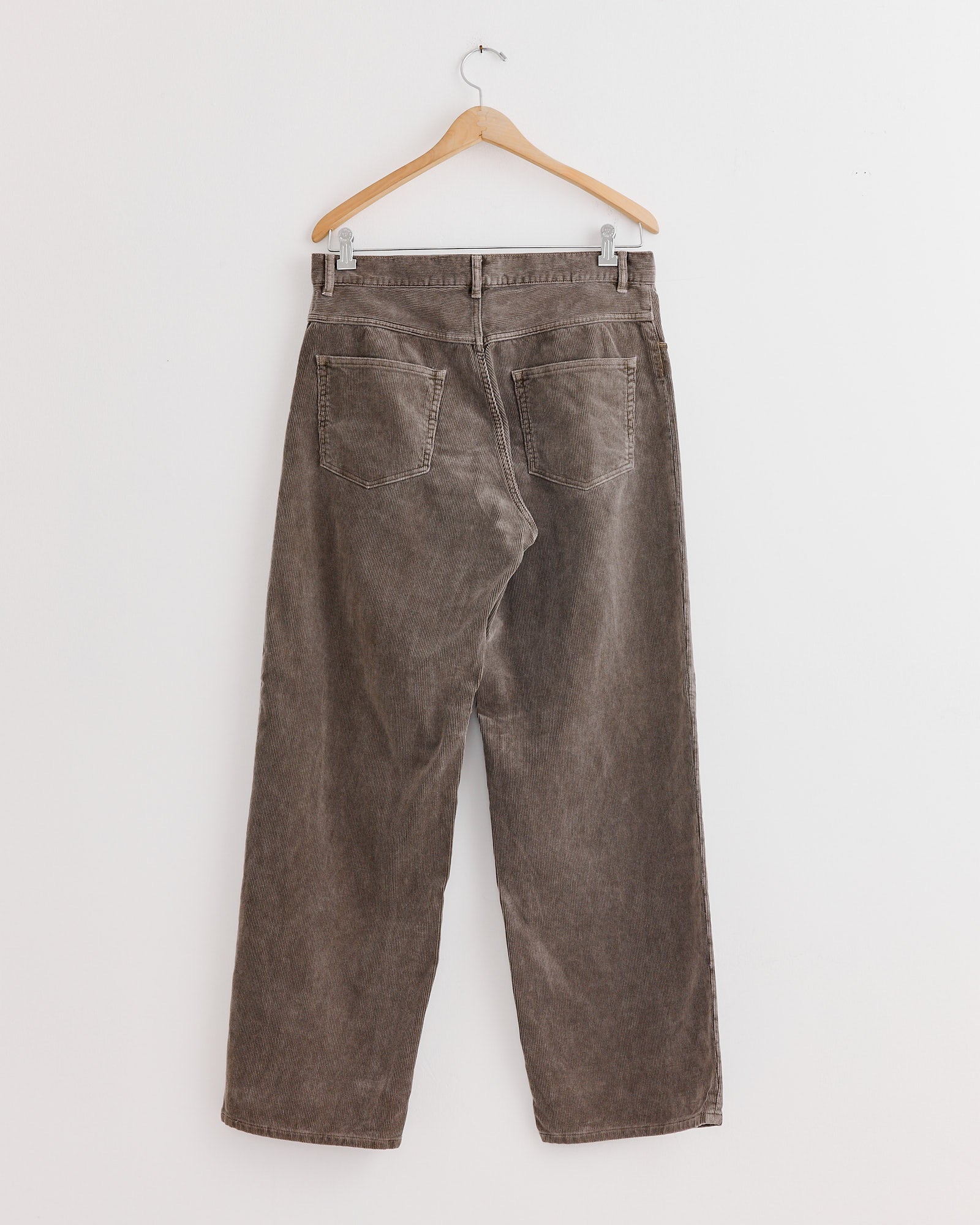 The Auralee Corduroy Wide 5 Pockets Pant in Olive Brown hangs on a wooden hanger against a white wall, shown from the back with rear pockets and belt loops—ideal for a flattering fit.