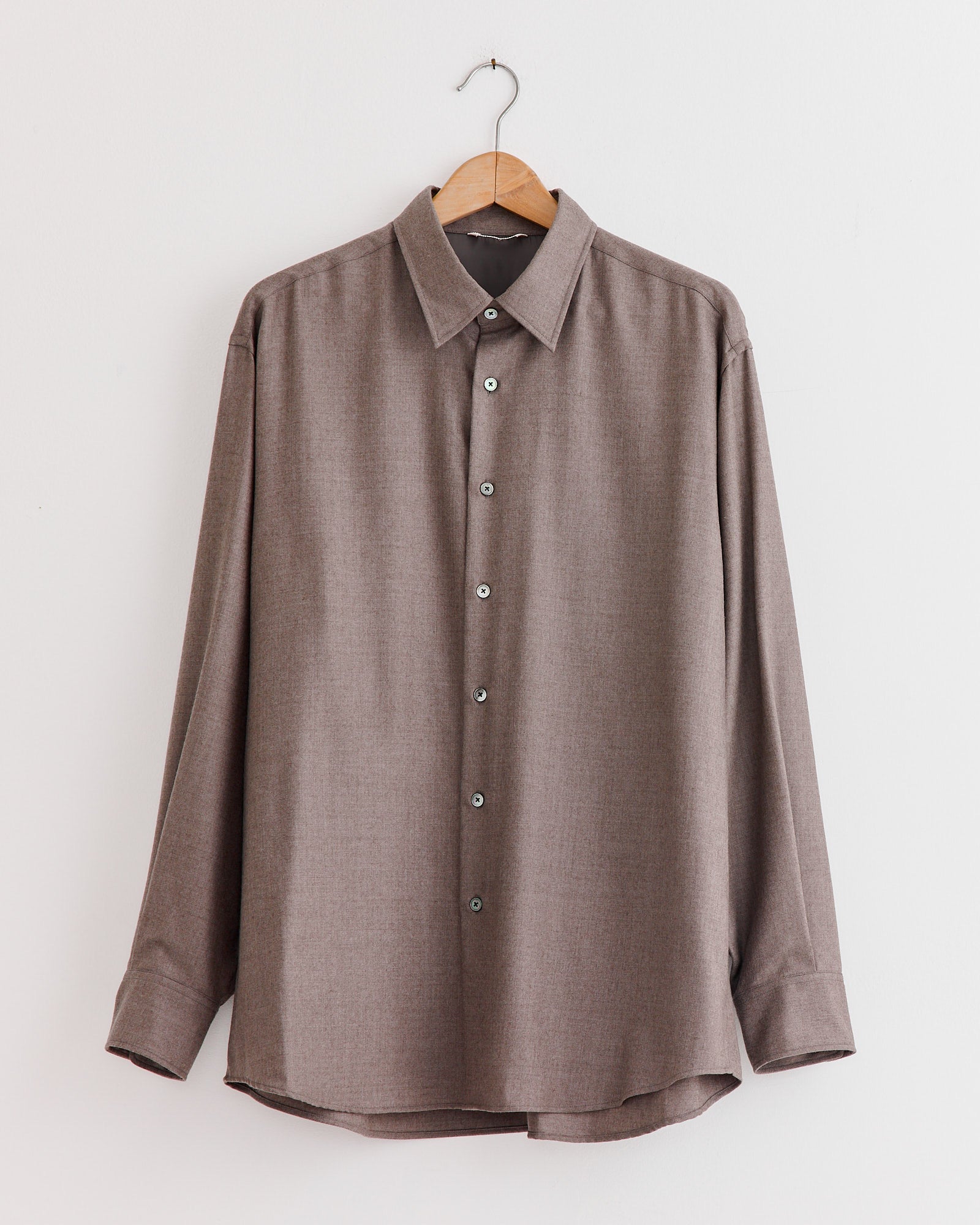 An Auralee Super Light Wool Shirt in Top Brown hangs on a wooden hanger against a plain white wall.